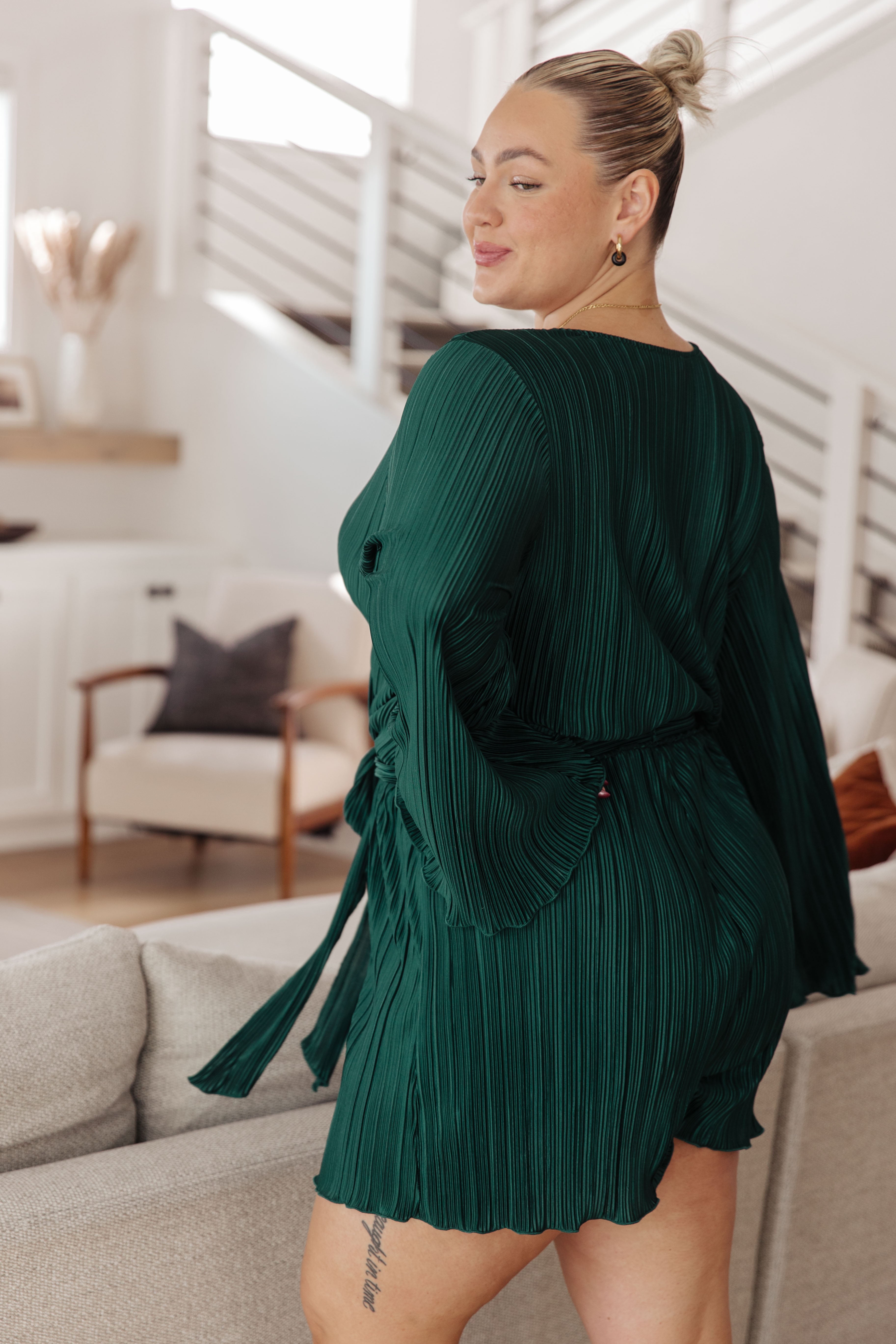 Plus-size woman with a blonde bun wearing a hunter green pleated romper with long sleeves and a tie waist, posing indoors by a light-colored couch.