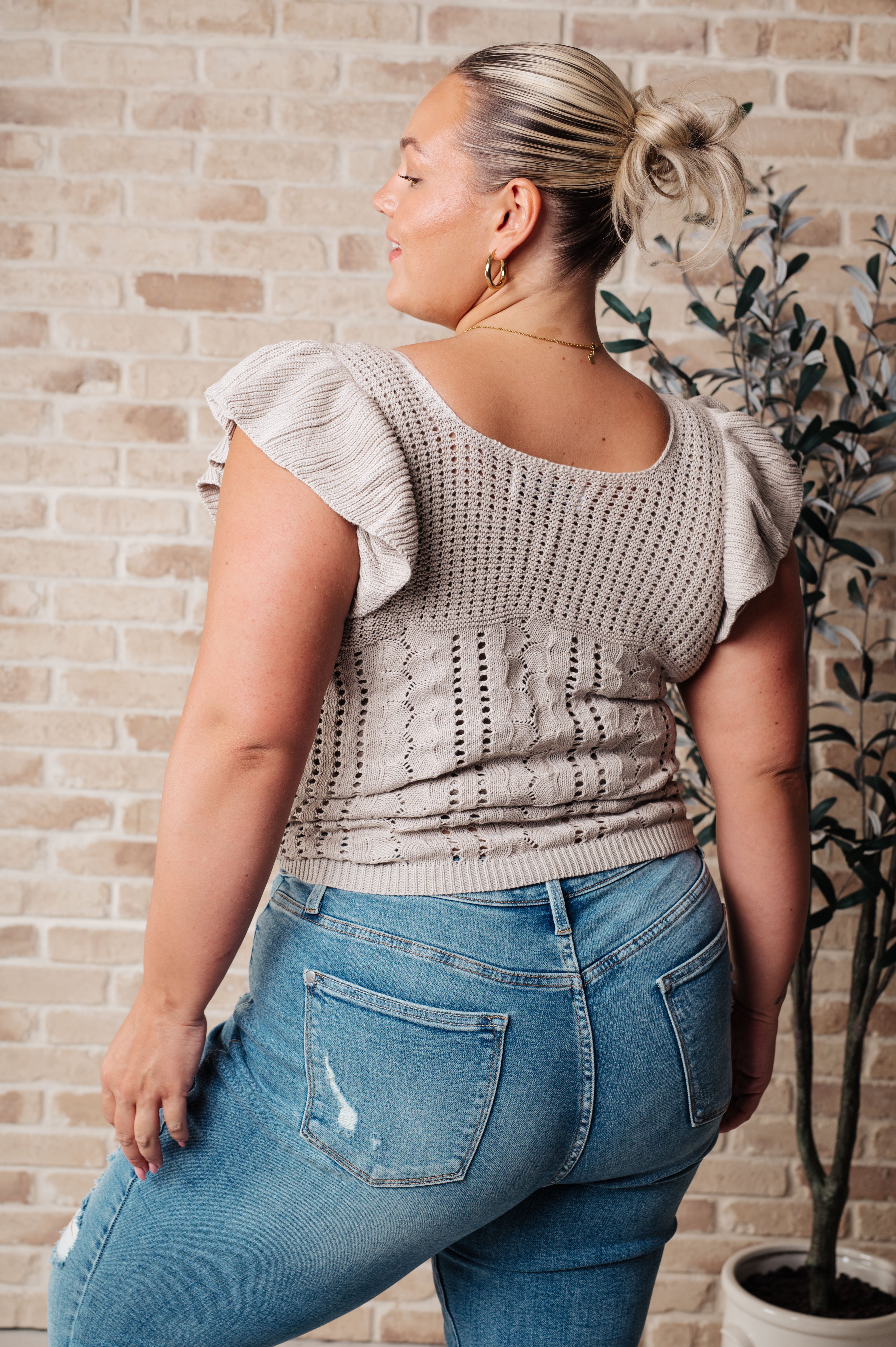 Curvy woman wearing a light grey pointelle knit top with ruffle sleeves paired with light blue distressed jeans, standing in front of a brick wall and plant.