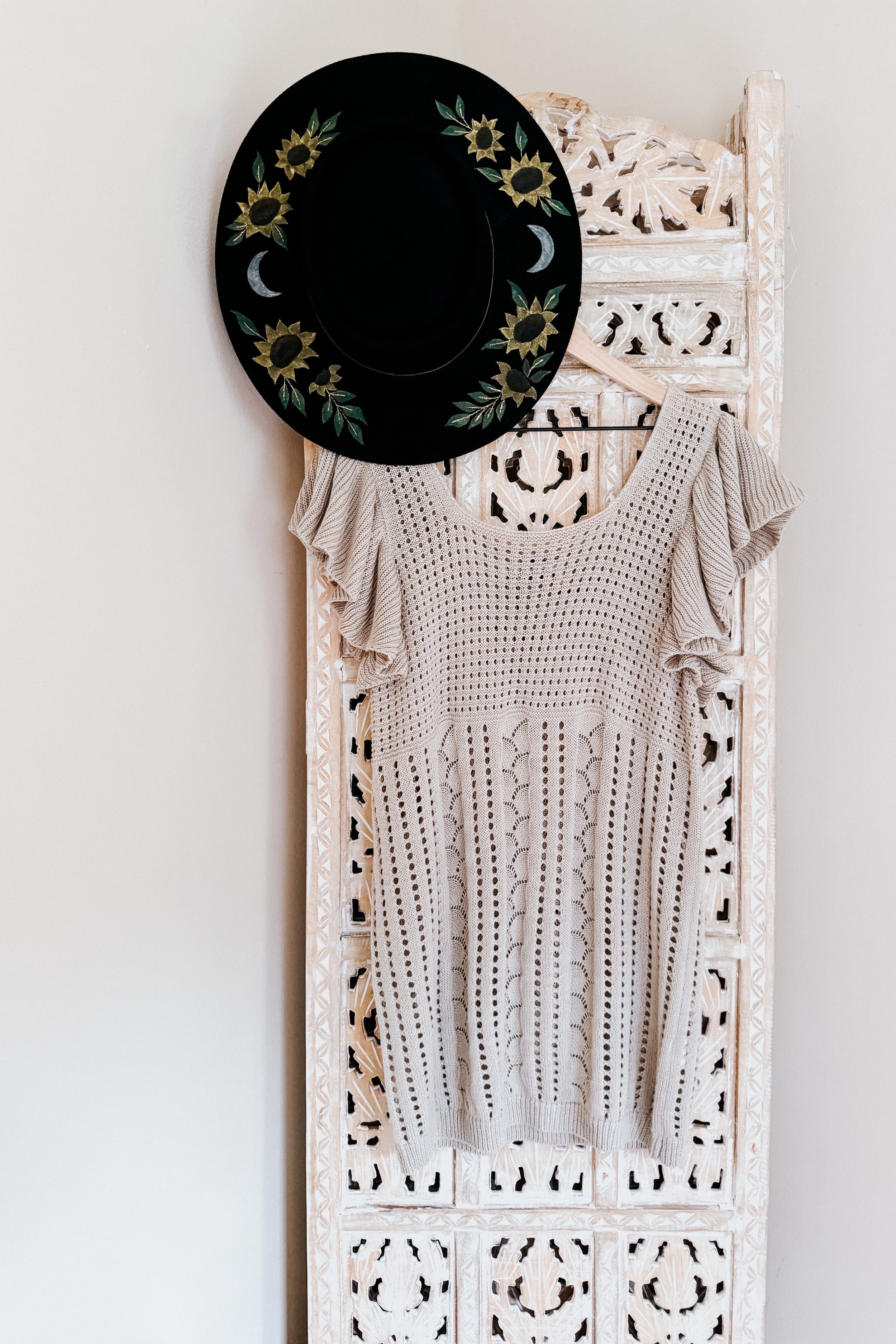Light grey pointelle knit top with flutter sleeves hanging on intricate carved white wooden panel next to black hat decorated with sunflower and moon embroidery
