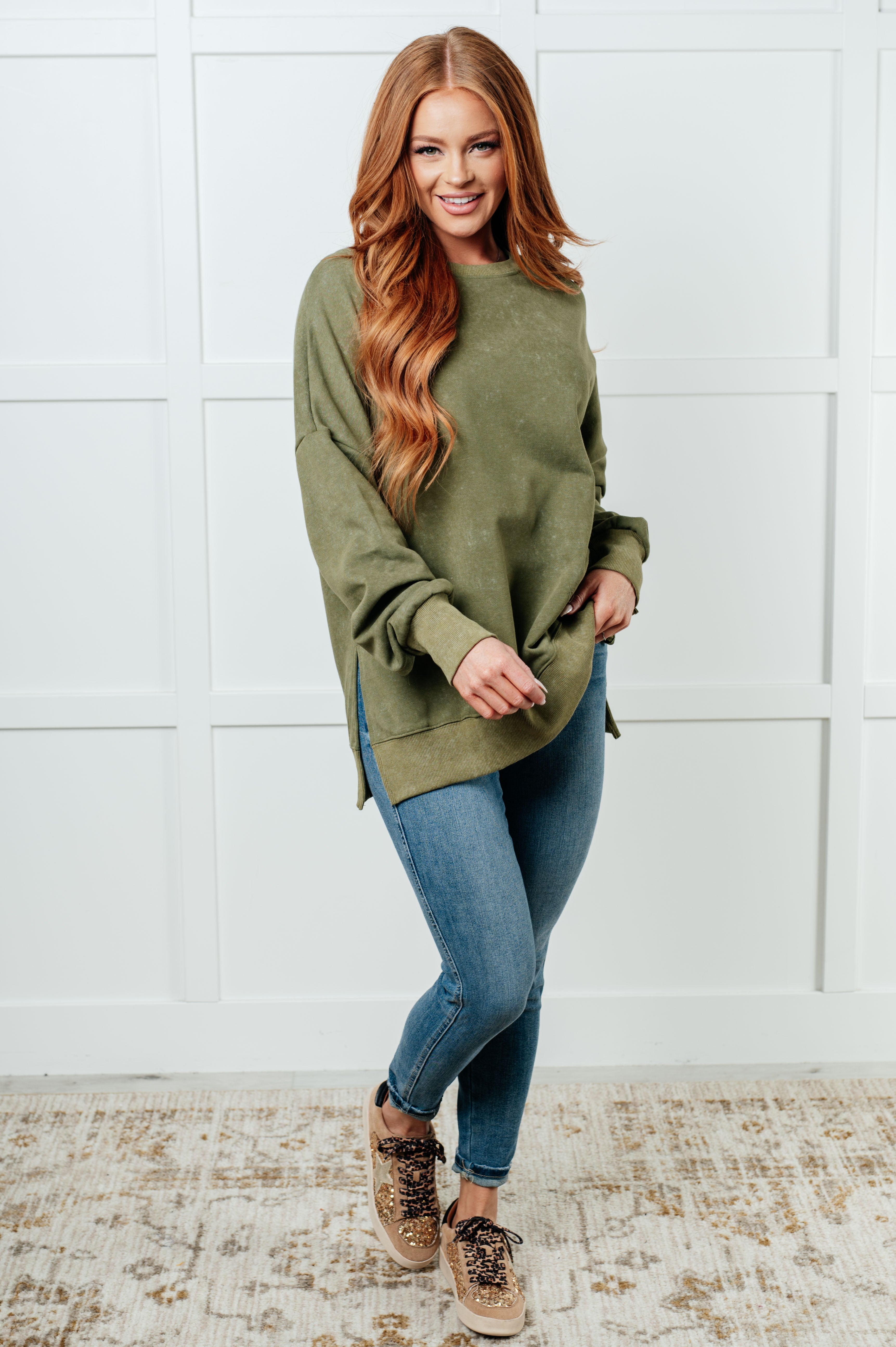 Smiling woman with long red hair wearing a green oversized sweatshirt paired with blue skinny jeans and leopard-print sneakers, standing indoors.