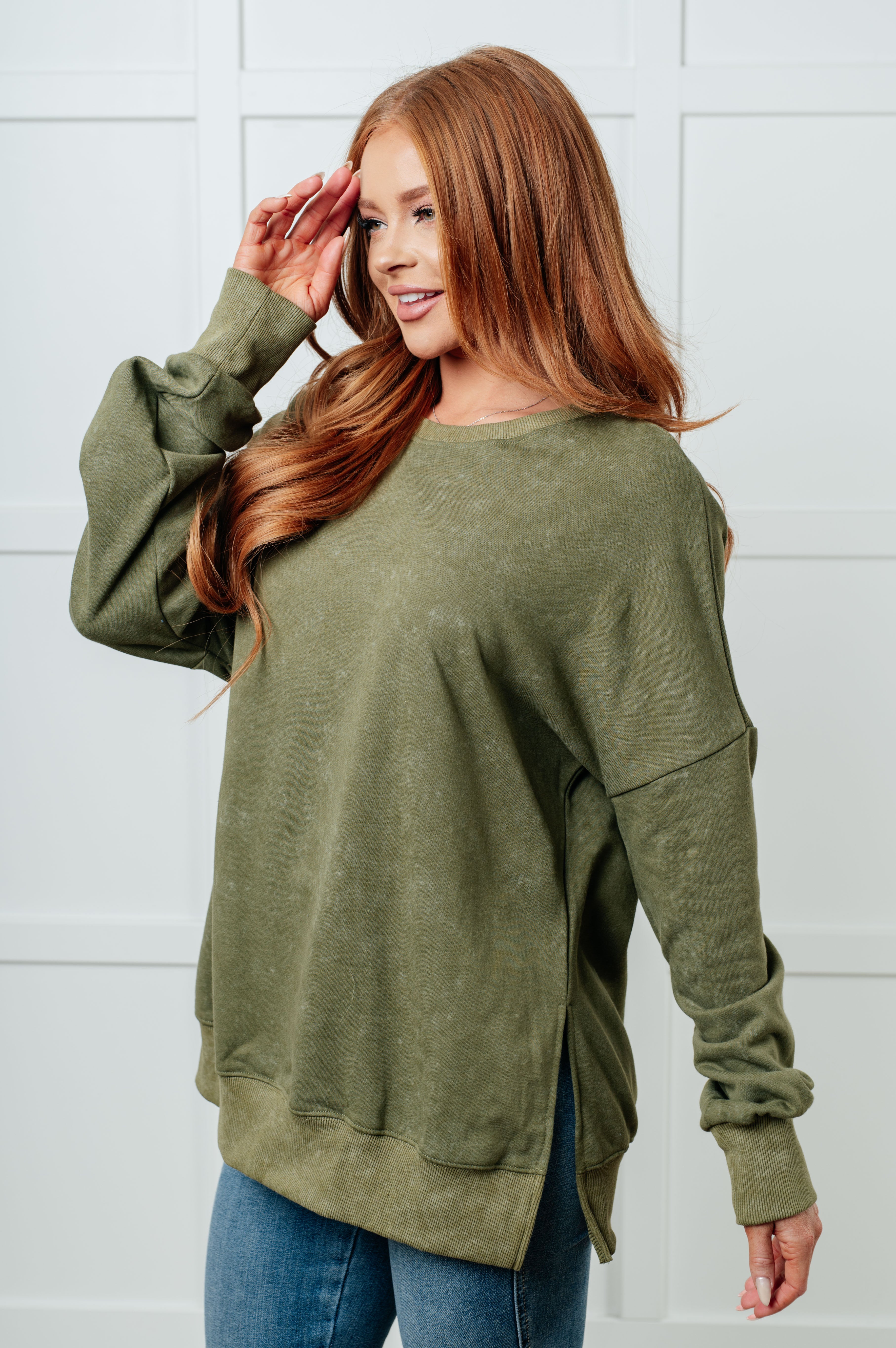 Red-haired woman wearing a relaxed green oversized sweatshirt with side slits paired with blue jeans, posing indoors against a white paneled wall.