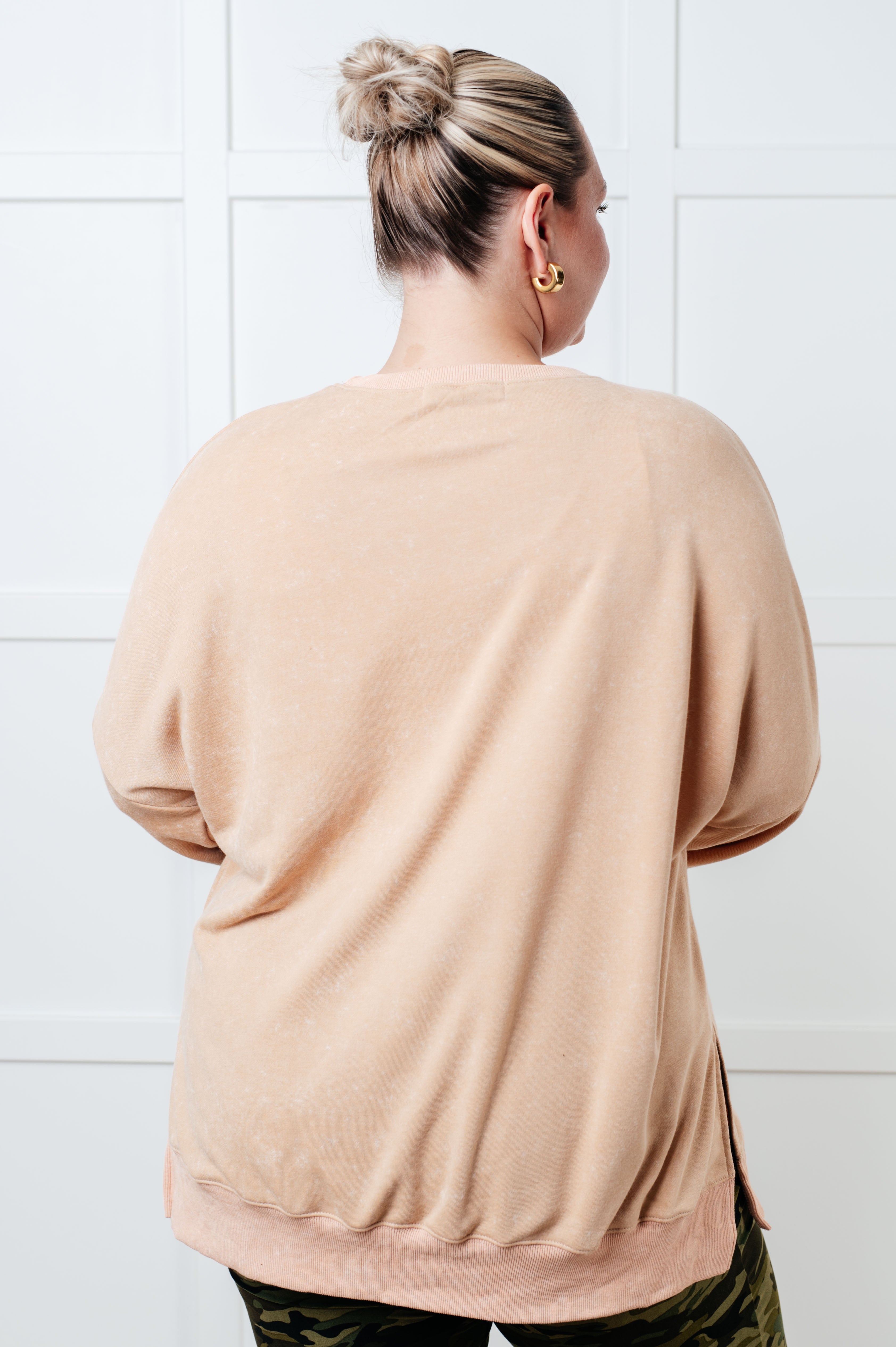 Woman with blonde hair in a bun wearing a relaxed khaki oversized sweatshirt shown from the back against a white paneled wall.