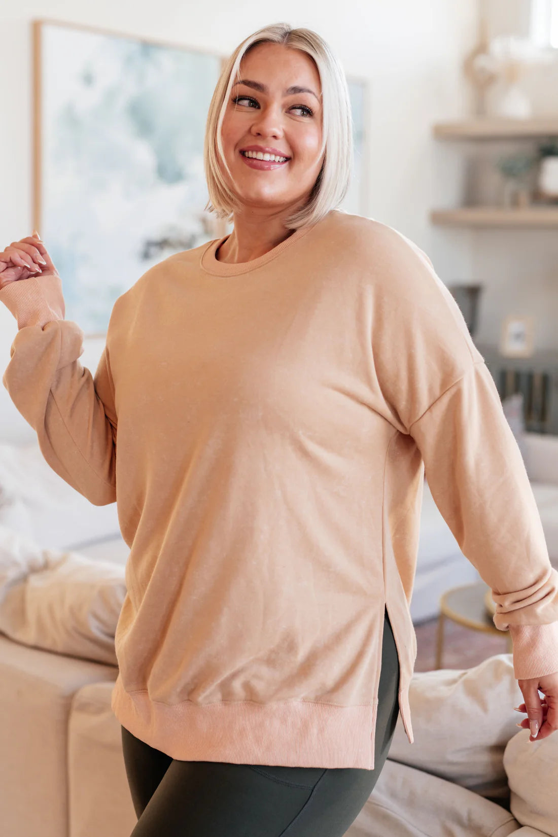 Smiling woman with blonde hair wearing a loose khaki oversized sweatshirt with side slits, standing in a bright cozy living room.
