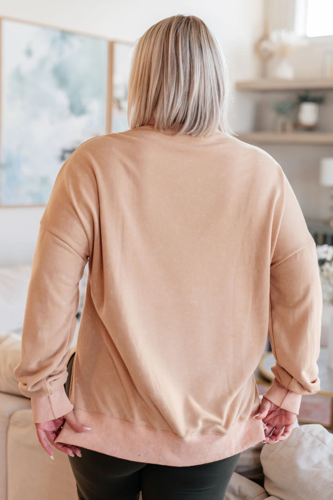 Woman with straight blonde hair wearing a loose-fitting khaki oversized sweatshirt, shown from behind in a cozy living room setting.
