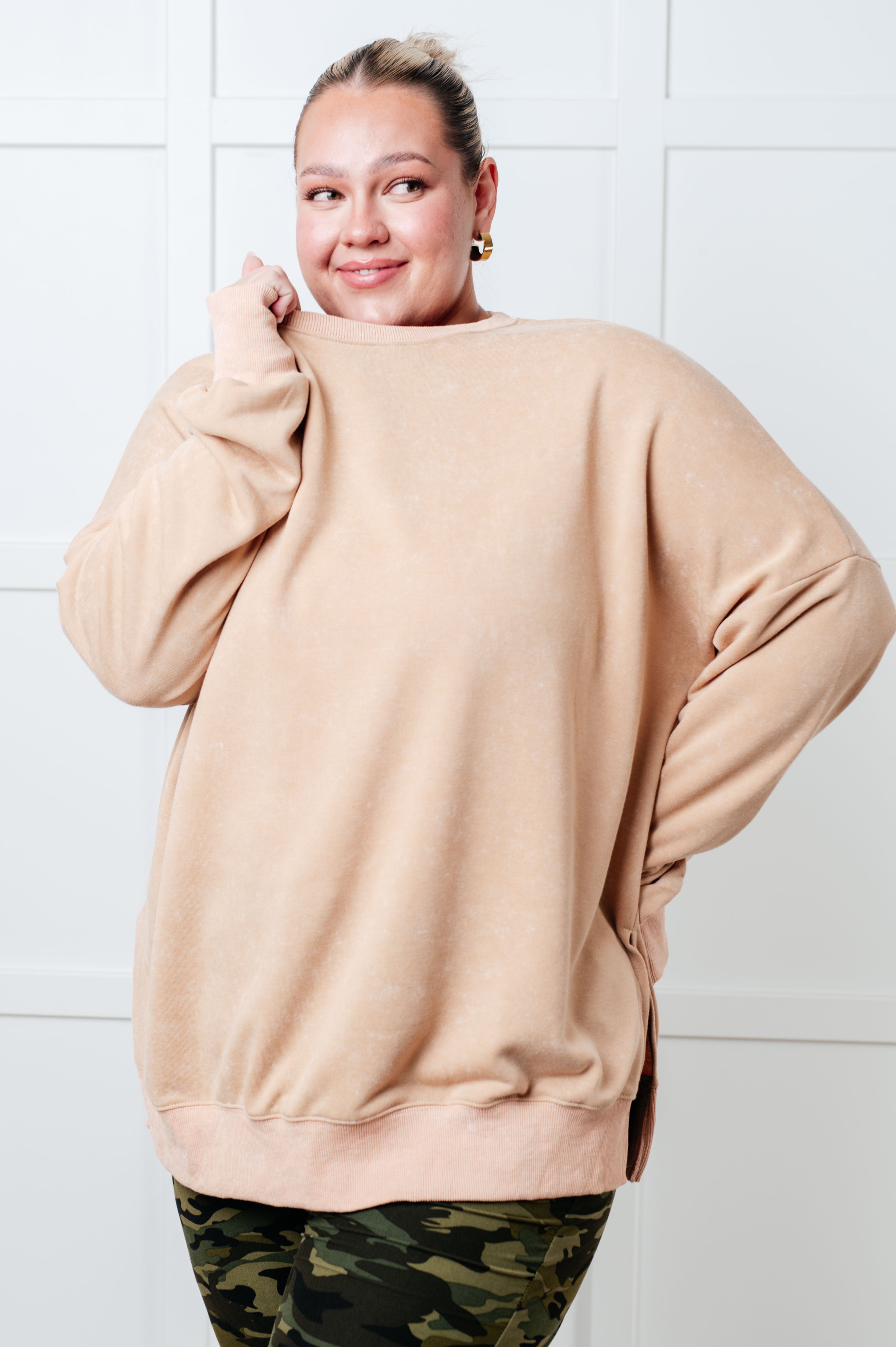 Smiling woman wearing a relaxed khaki oversized sweatshirt with side slits paired with camo leggings, posed against a white paneled wall.