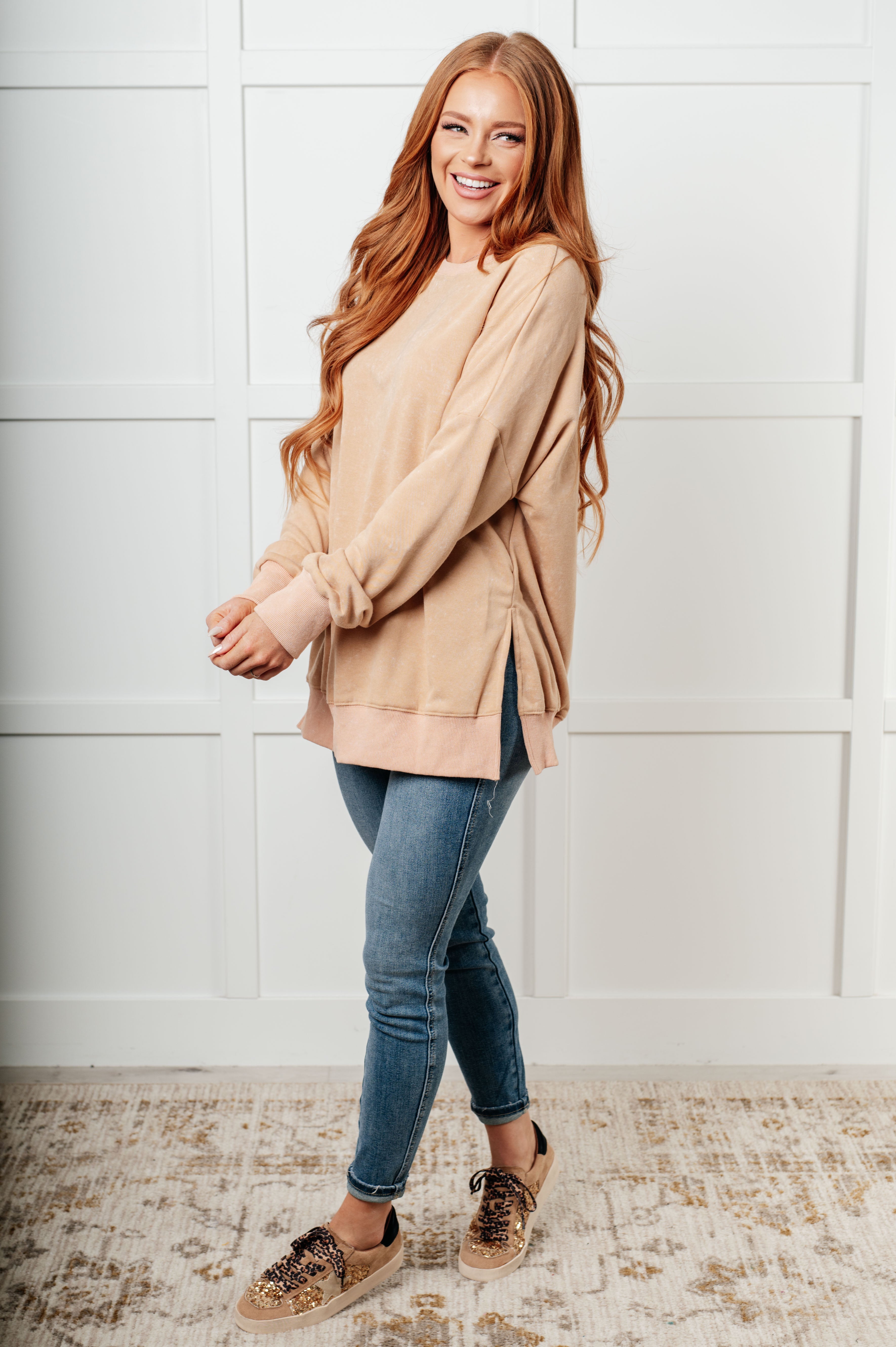 Woman with long red hair wearing a relaxed khaki oversized sweatshirt with side slits paired with slim blue jeans and leopard-print sneakers, smiling indoors.