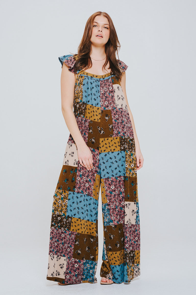 Woman with long brown hair wearing a relaxed patchwork jumpsuit with flutter sleeves in brown, blue, and mustard tones against a plain background.