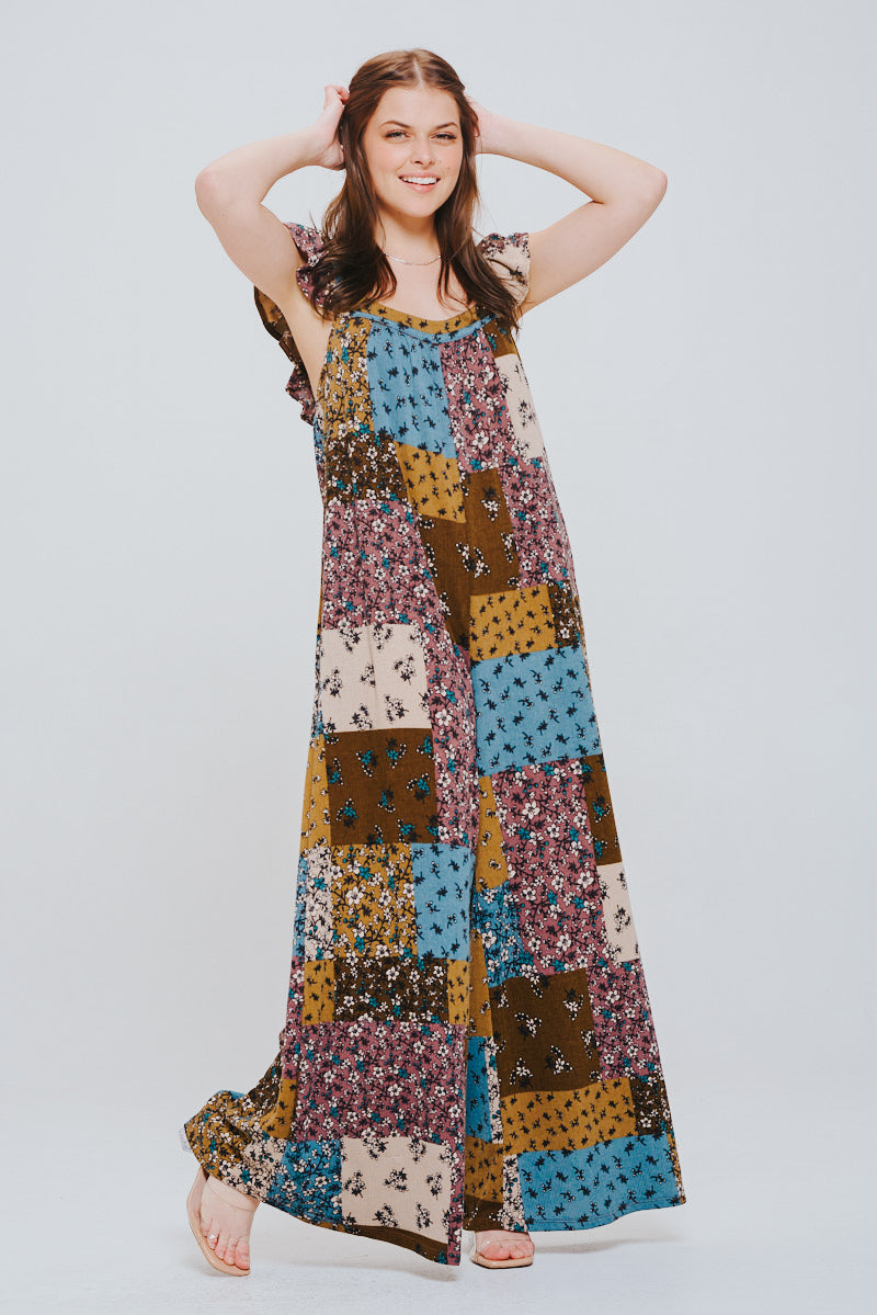Smiling woman with long brown hair wearing a loose-fitting patchwork knit jumpsuit in brown, blue, pink, and mustard tones with ruffle sleeves against a plain background.