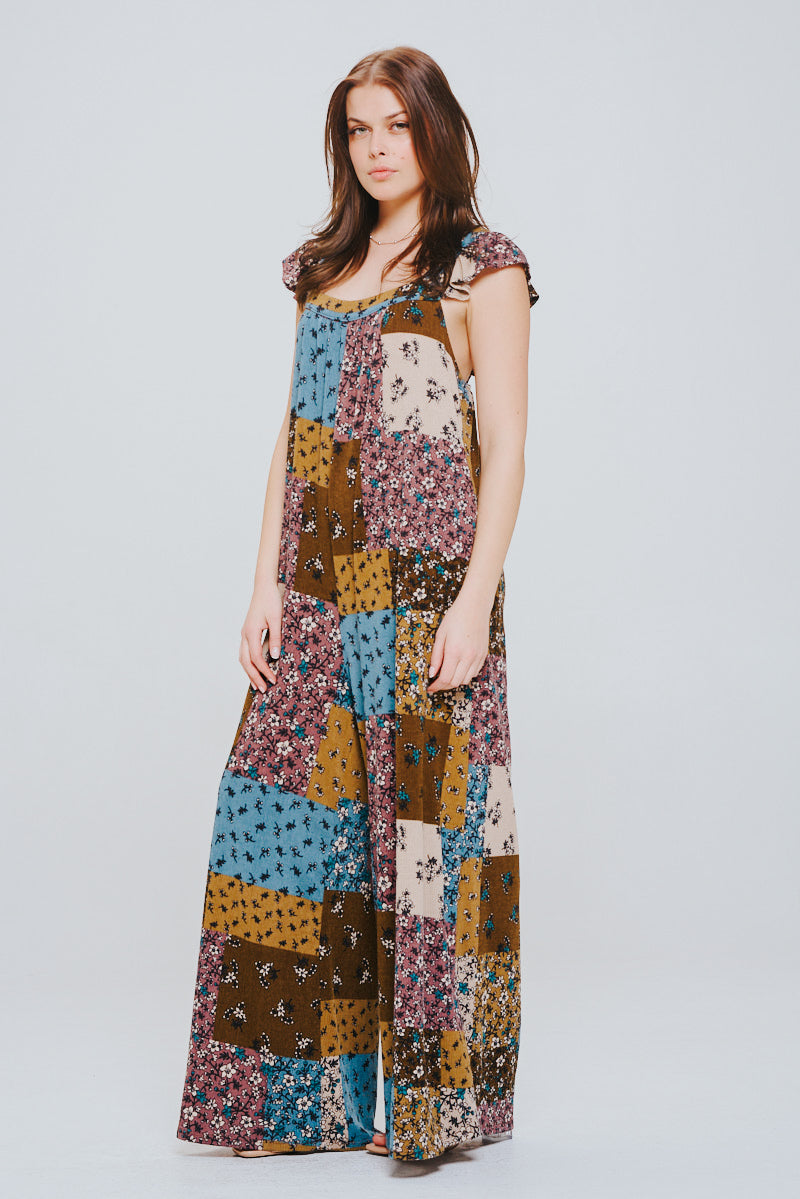 Young woman with brown hair wearing a sleeveless patchwork maxi jumpsuit in brown, blue, mustard, and cream floral prints against a plain background.
