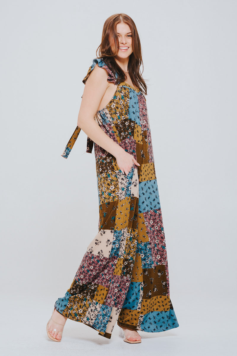 Smiling woman wearing a sleeveless patchwork jumpsuit in brown, blue, mustard, and cream tones with ruffle straps, standing with one hand in pocket.