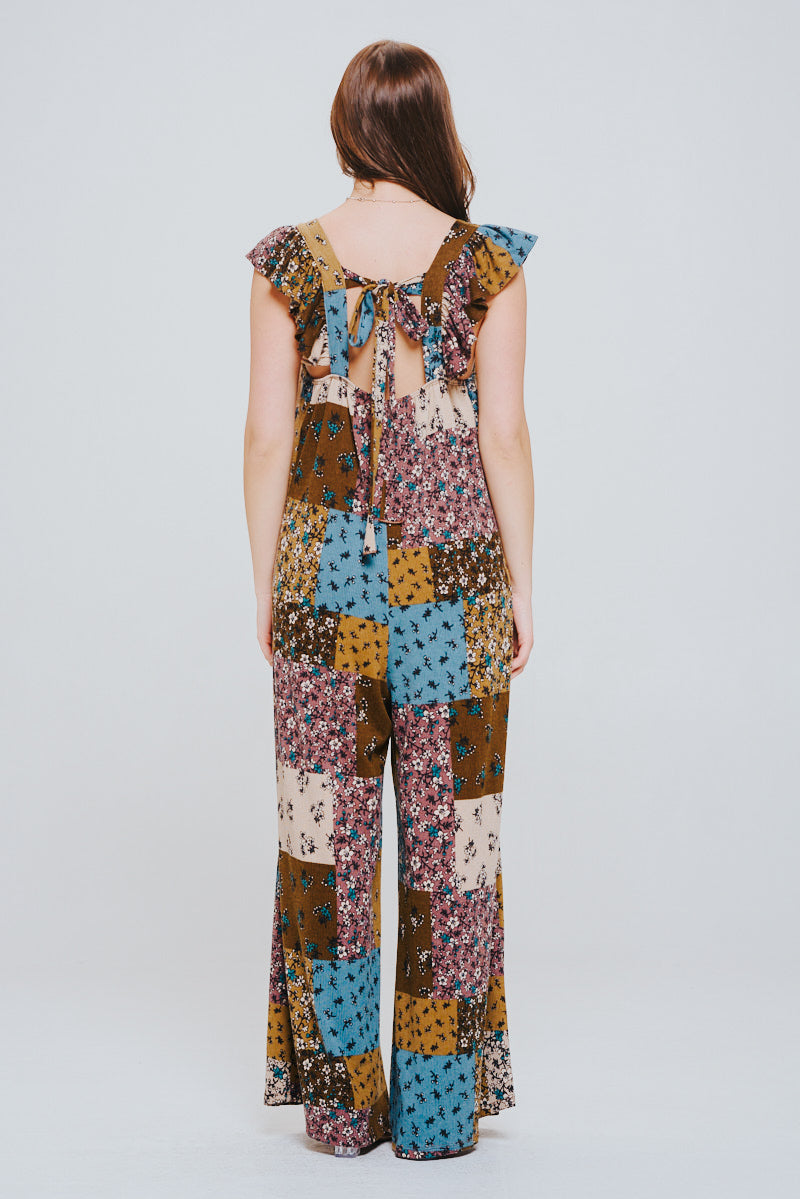 Woman with long brown hair shown from the back wearing a sleeveless patchwork knit jumpsuit in brown, blue, mustard, and cream tones with ruffle sleeves and an open back tied with bows.