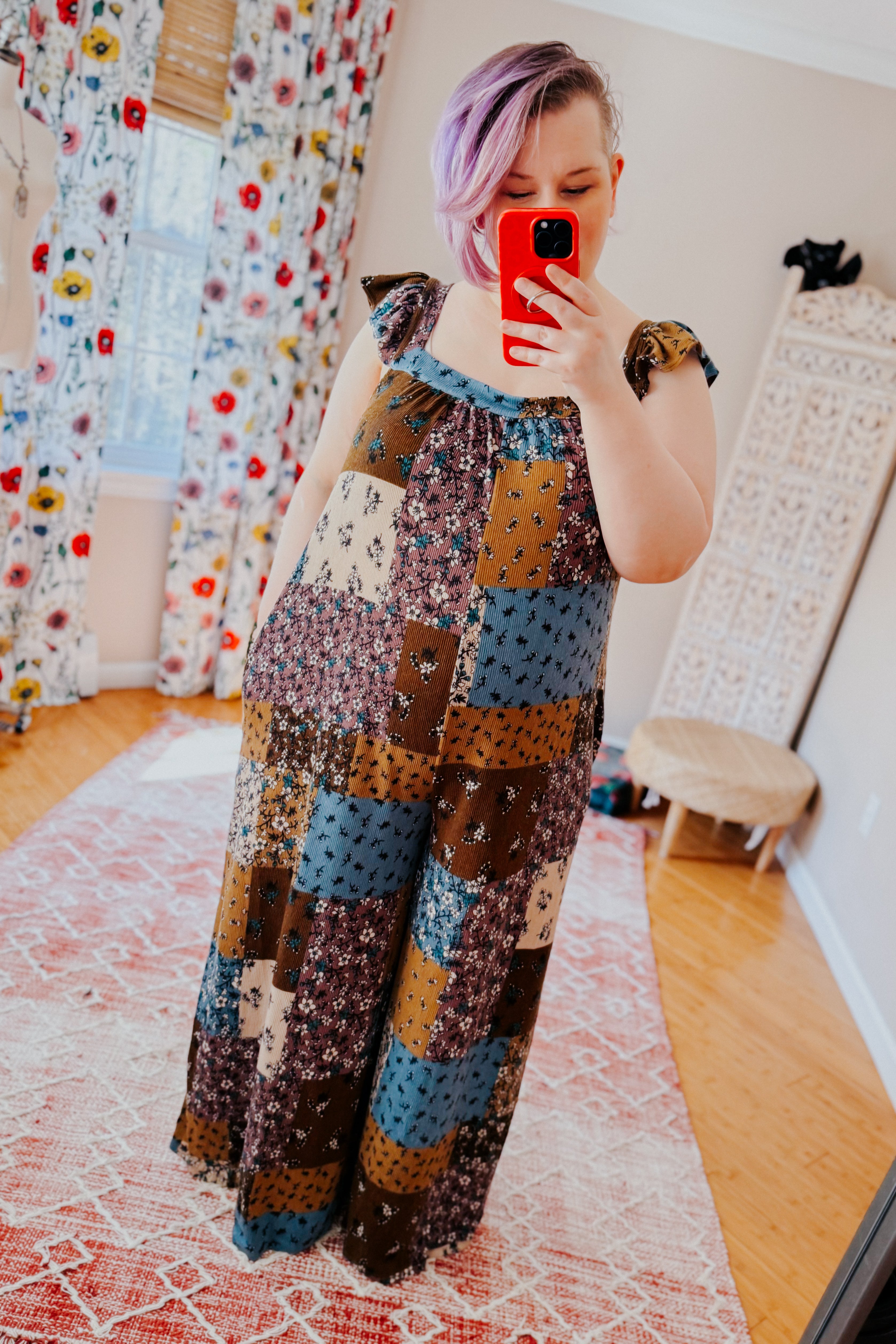 Woman with short light purple hair taking a mirror selfie wearing a loose-fitting knit patchwork jumpsuit in brown, blue, and cream tones with ruffle sleeve details.