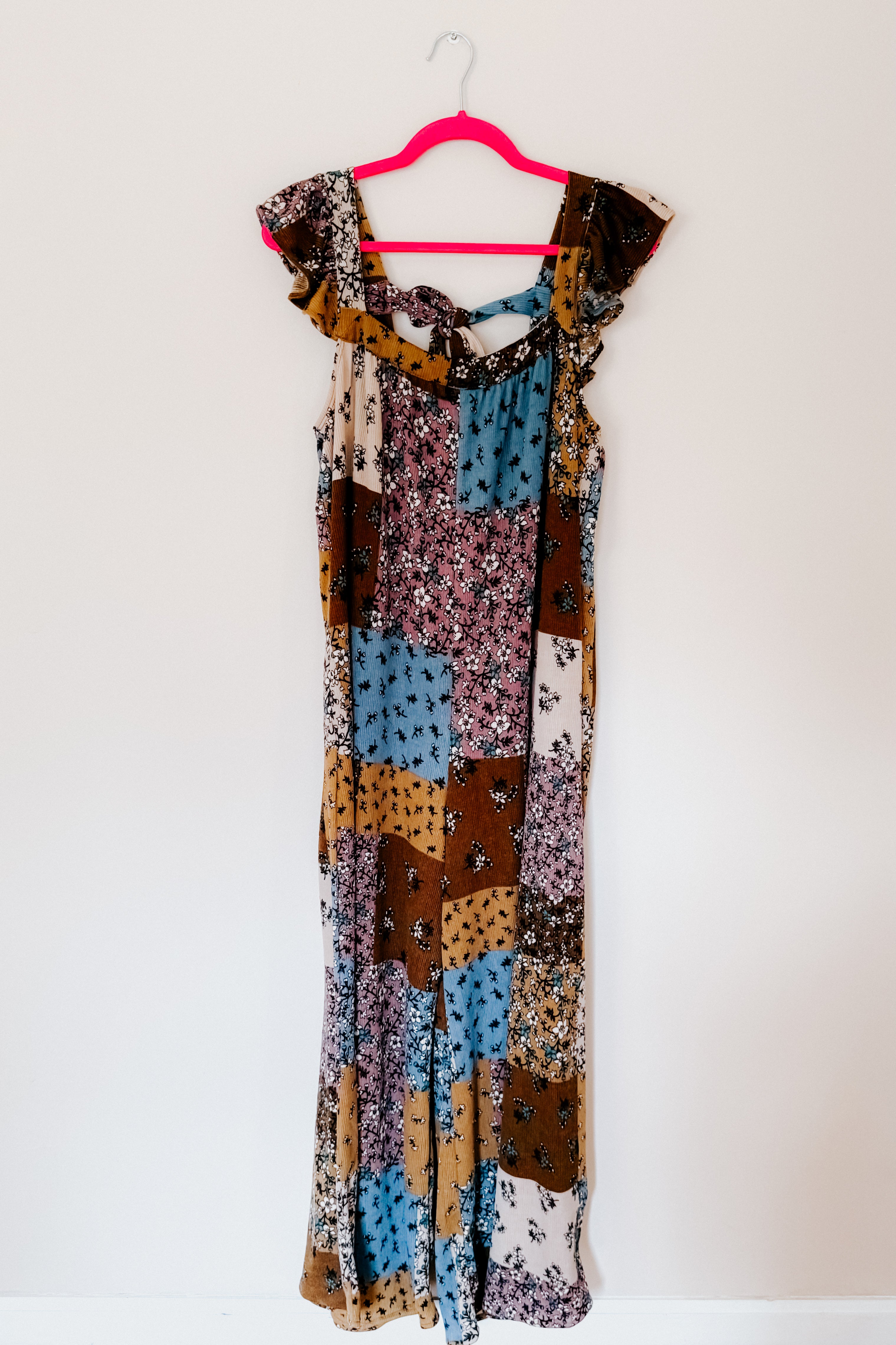 Sleeveless knit jumpsuit with ruffle trim in brown, blue, cream, and mustard patchwork featuring floral patterns, displayed on a pink hanger.