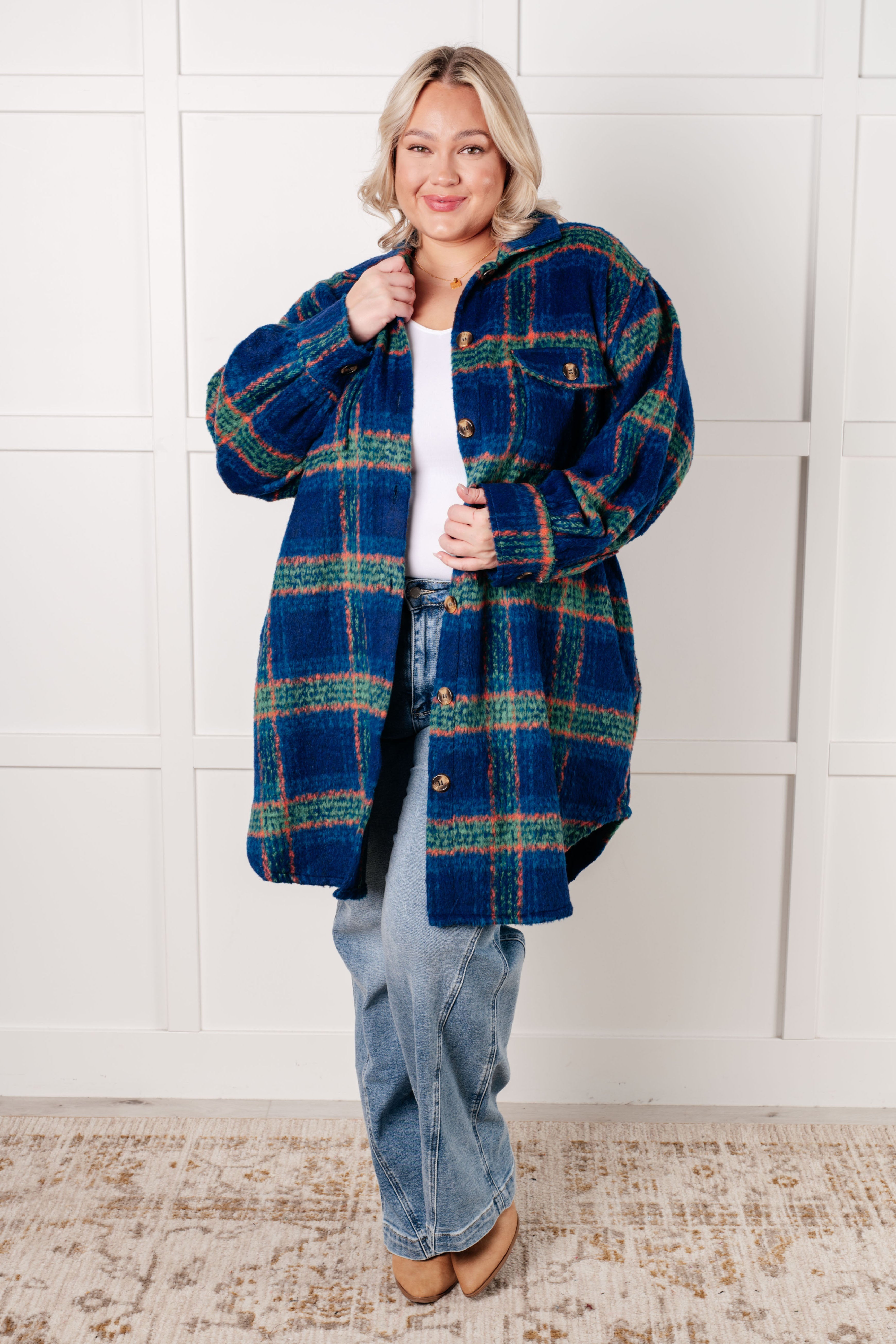 Smiling woman wearing a cozy blue plaid coat with green and orange accents over a white top and light washed jeans against a white paneled wall.
