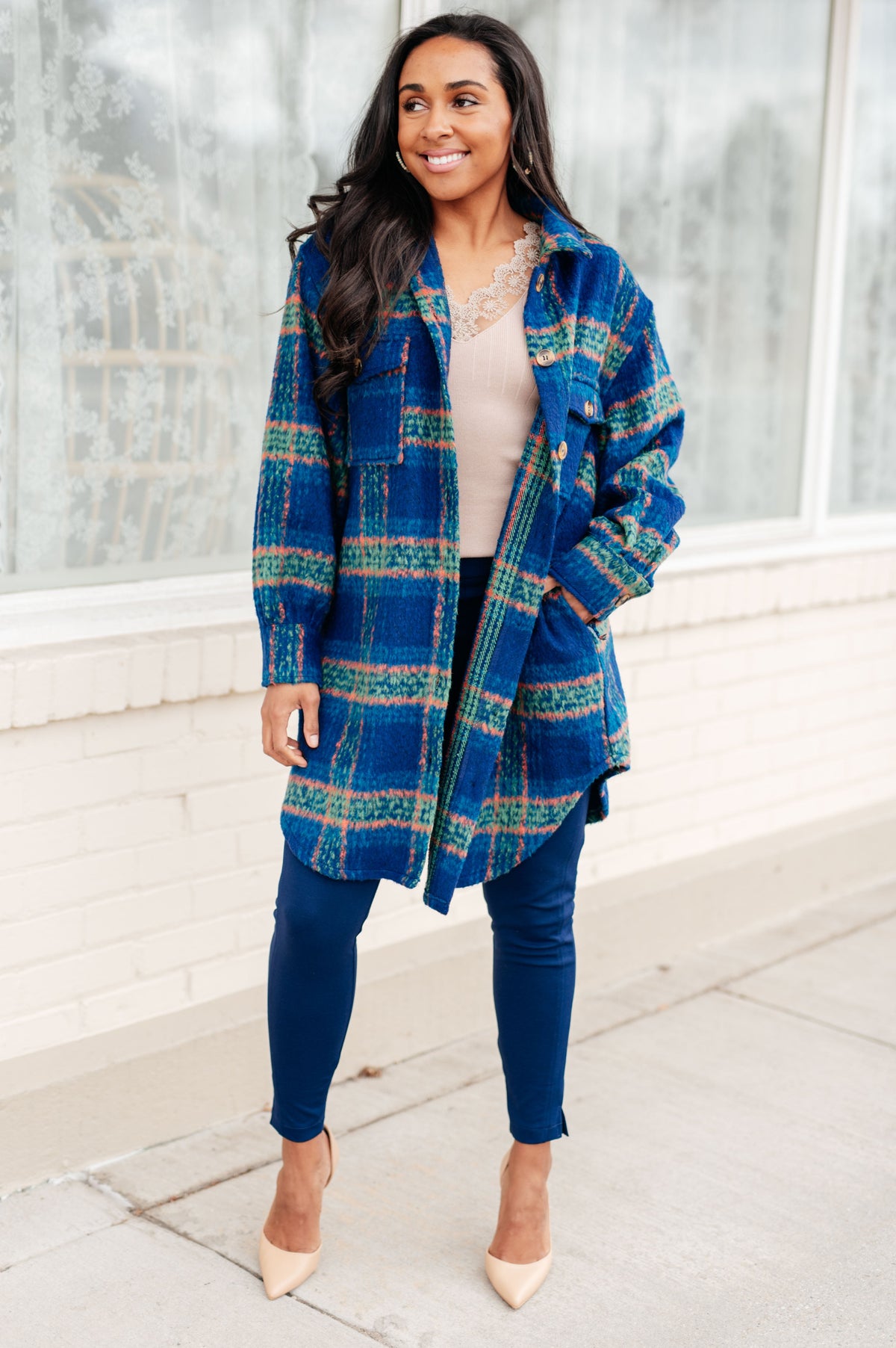 Woman modeling a blue plaid button front coat with a scooped hem over a lace top with dark leggings and nude heels indoors