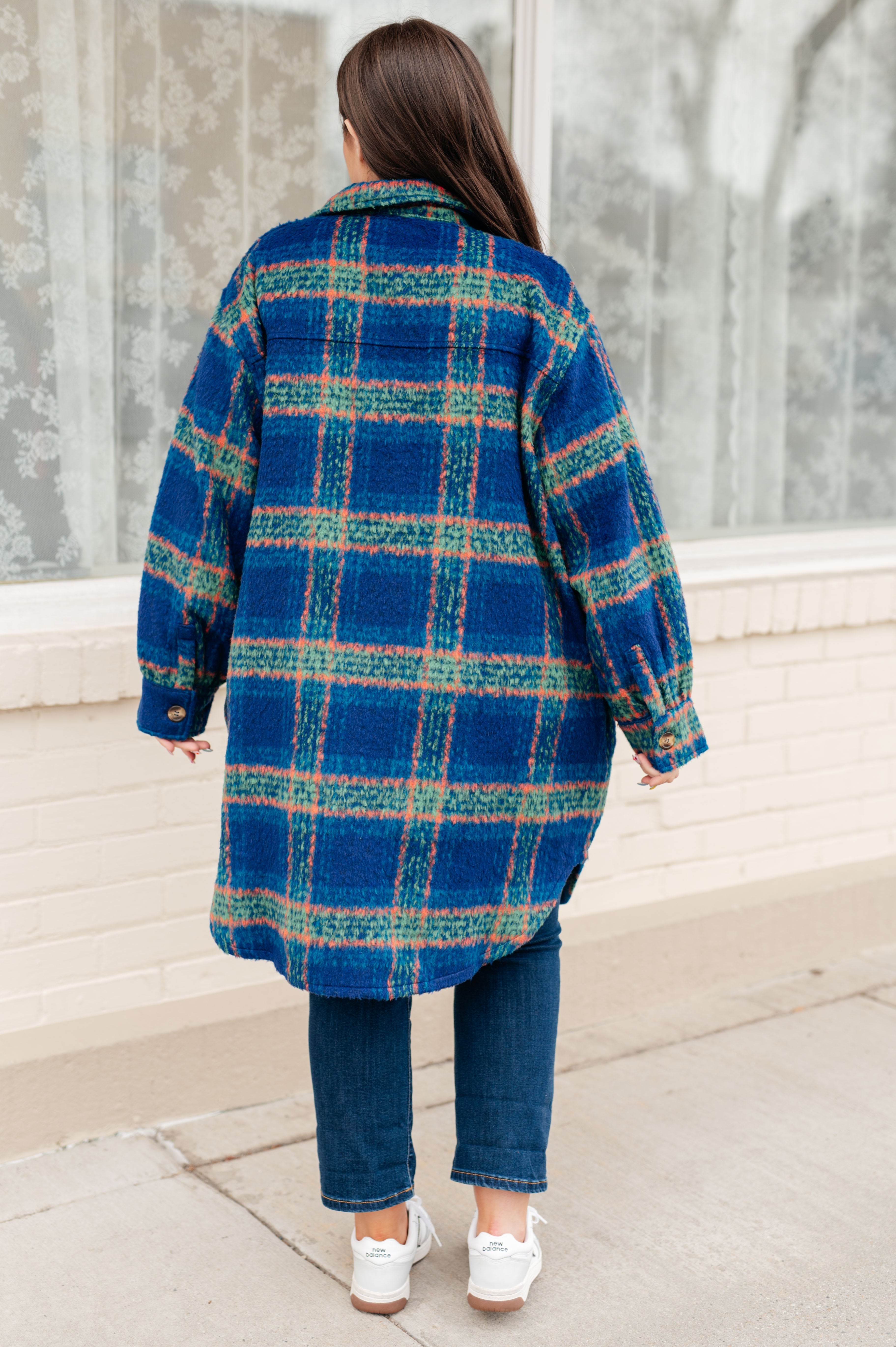 Woman wearing a blue plaid coat with green and coral accents over dark jeans and white sneakers, standing outdoors with her back to the camera.