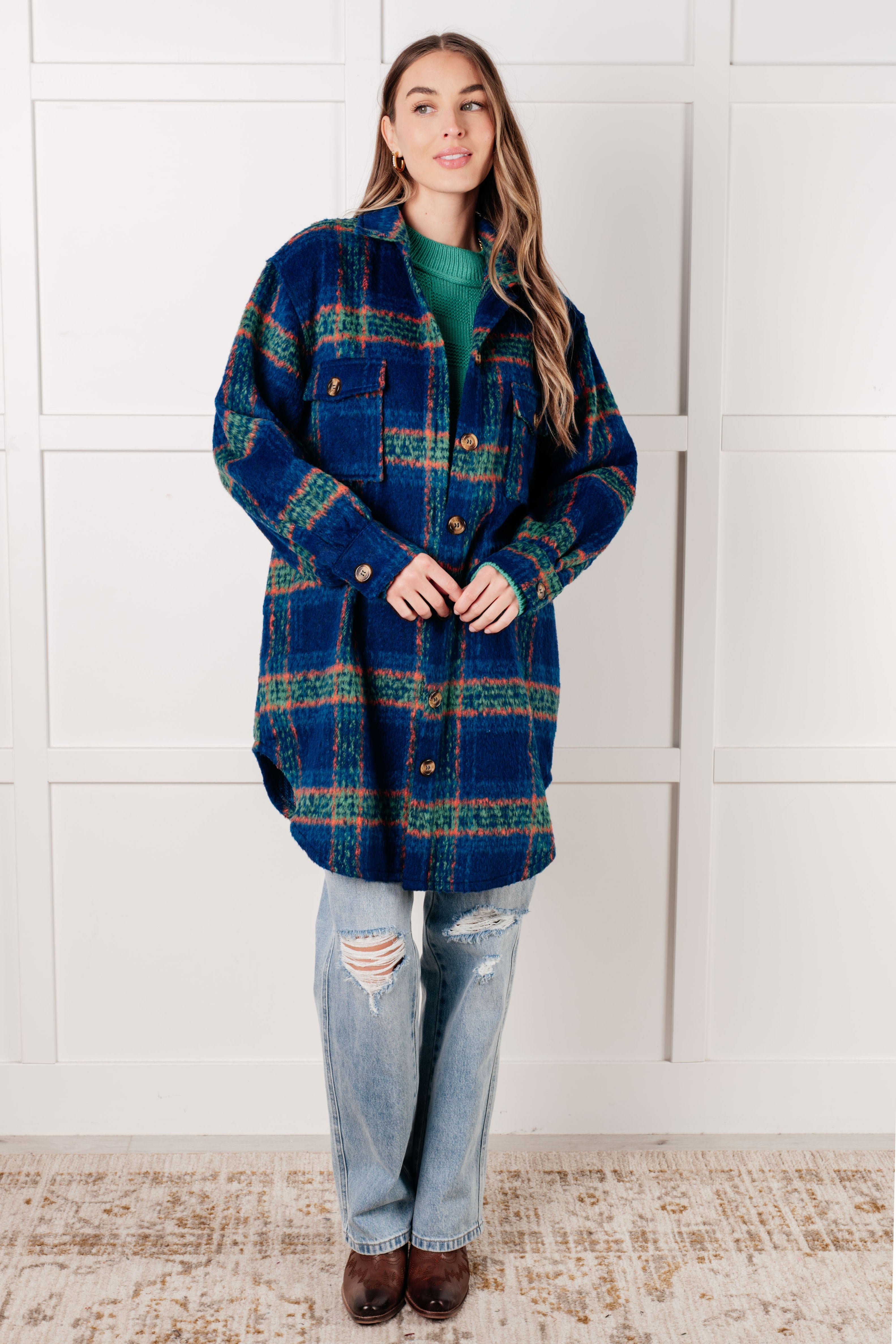 Woman wearing a blue plaid shacket with green and red accents over a green sweater, paired with ripped light-wash jeans and brown boots against a white paneled wall.