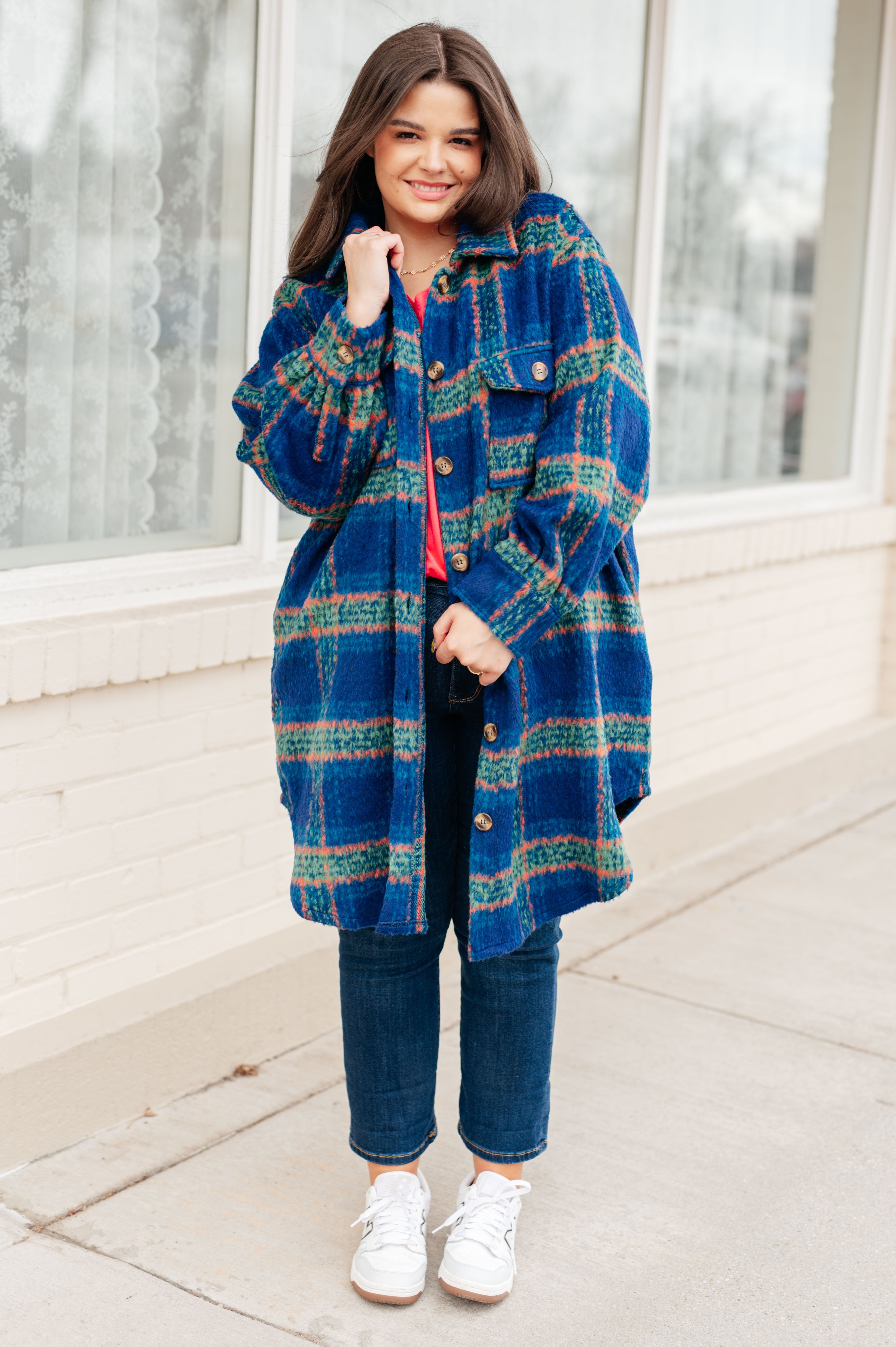 Smiling woman wearing a cozy blue plaid coat with green and peach stripes over a red top and dark jeans, standing outside.