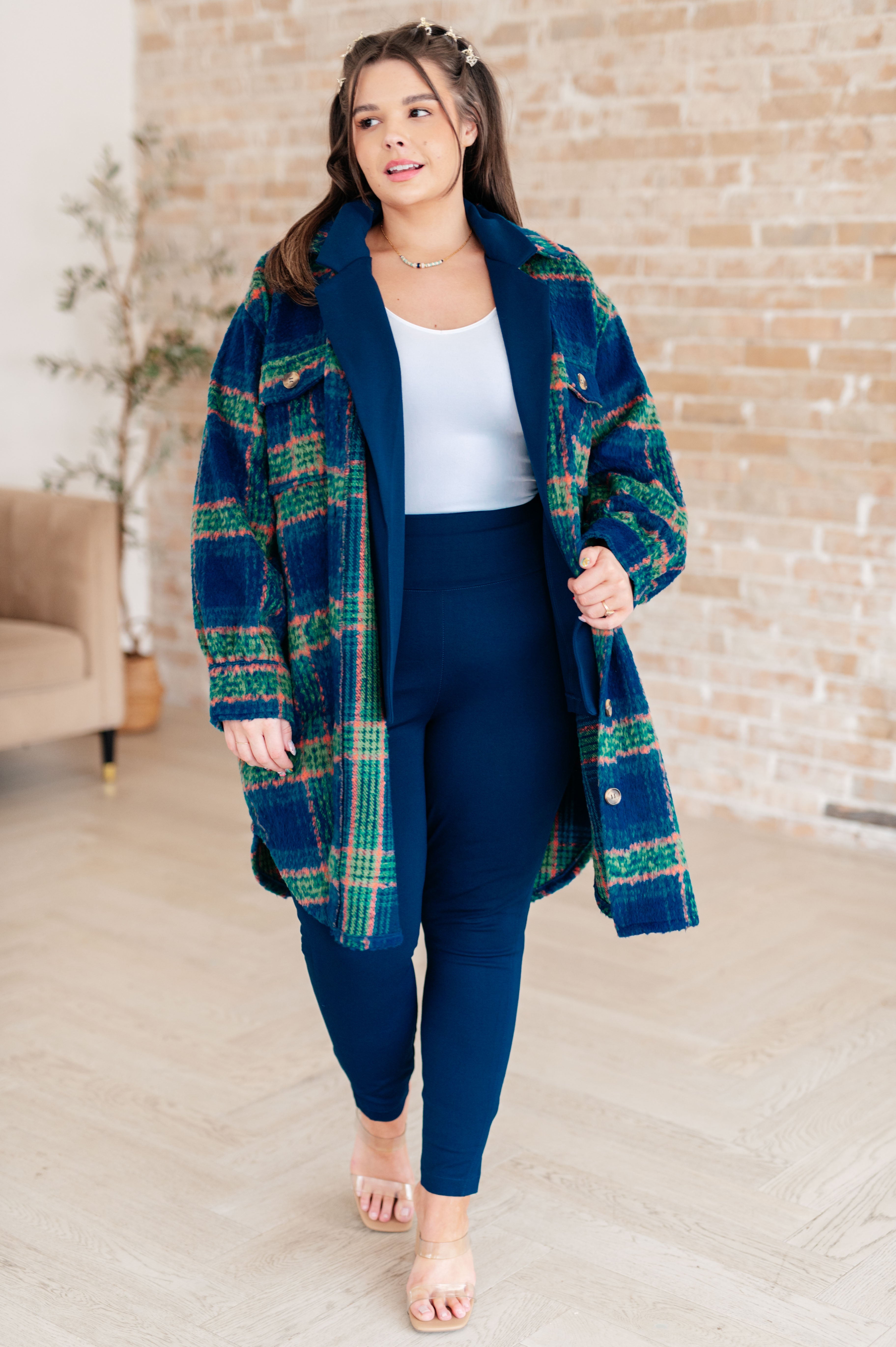Woman wearing a blue plaid coat with green and orange accents paired with navy leggings and a white tank top walking indoors against a brick wall backdrop.