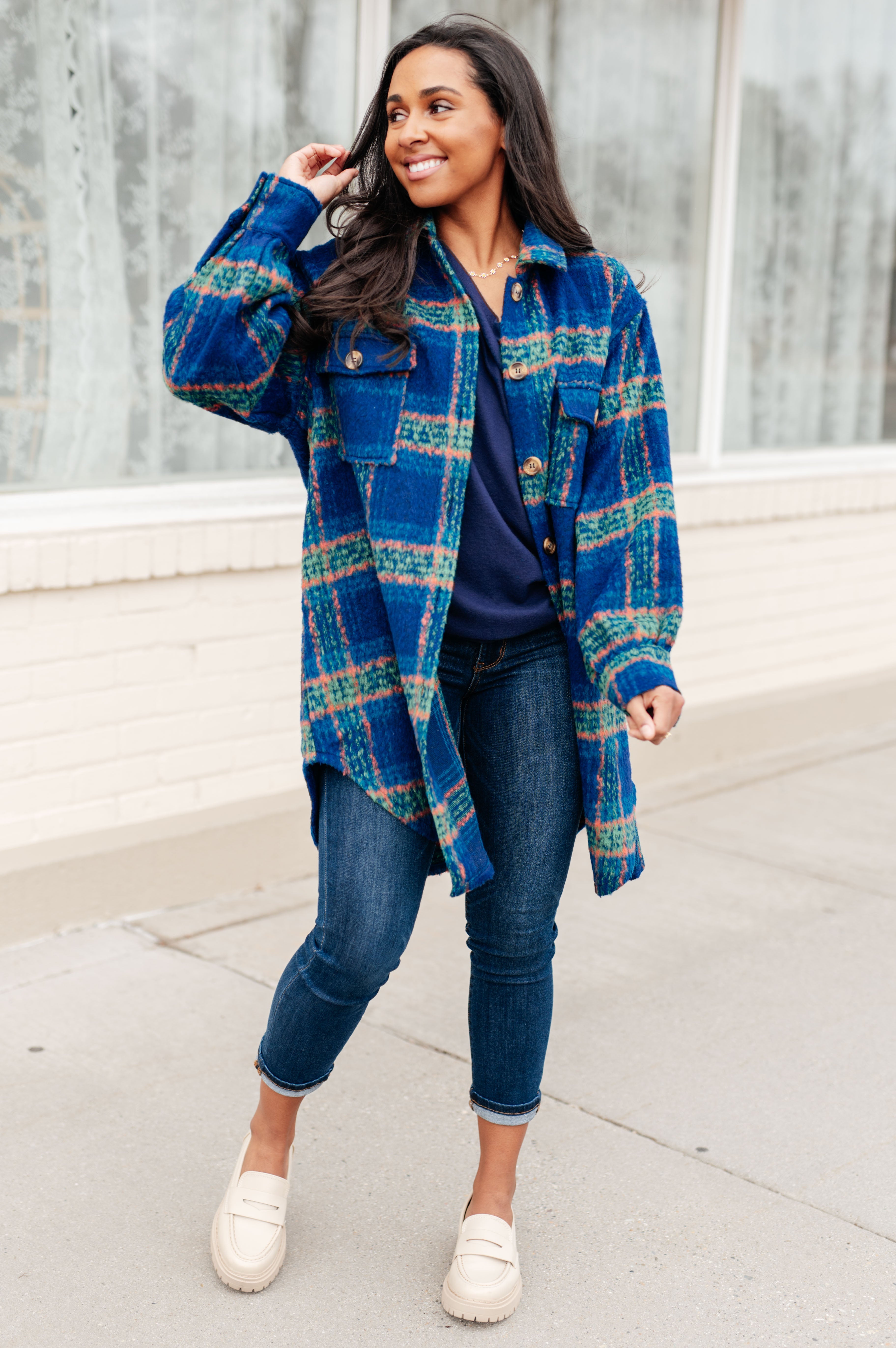 Woman smiling wearing a blue plaid overshirt with green and peach stripes, paired with dark jeans and cream loafers, standing outdoors.