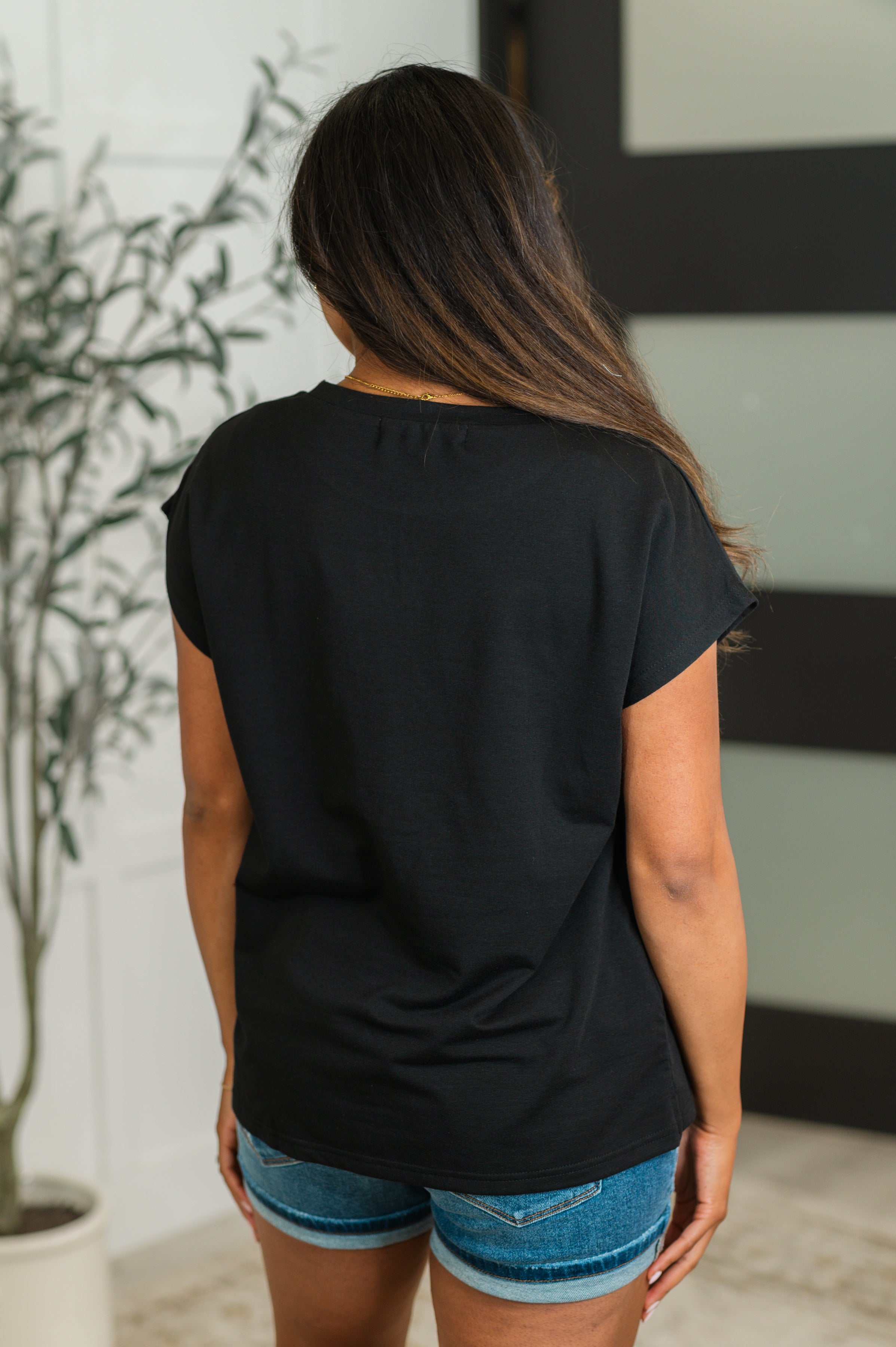 Woman shown from behind wearing a relaxed black short-sleeve tee paired with denim shorts in a casual indoor setting.