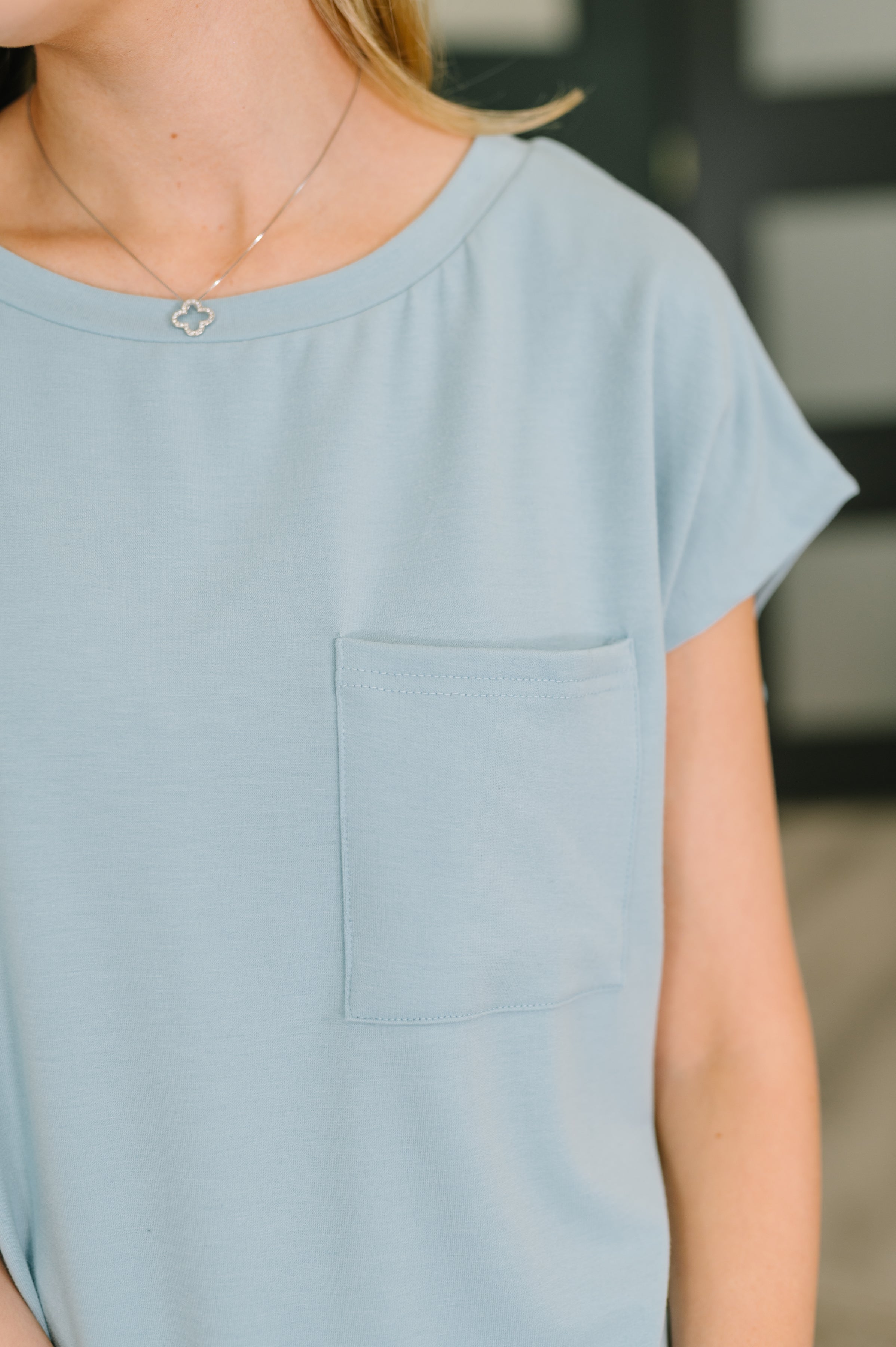 Close-up of soft blue short-sleeve tee with chest pocket and delicate silver necklace against blurred background