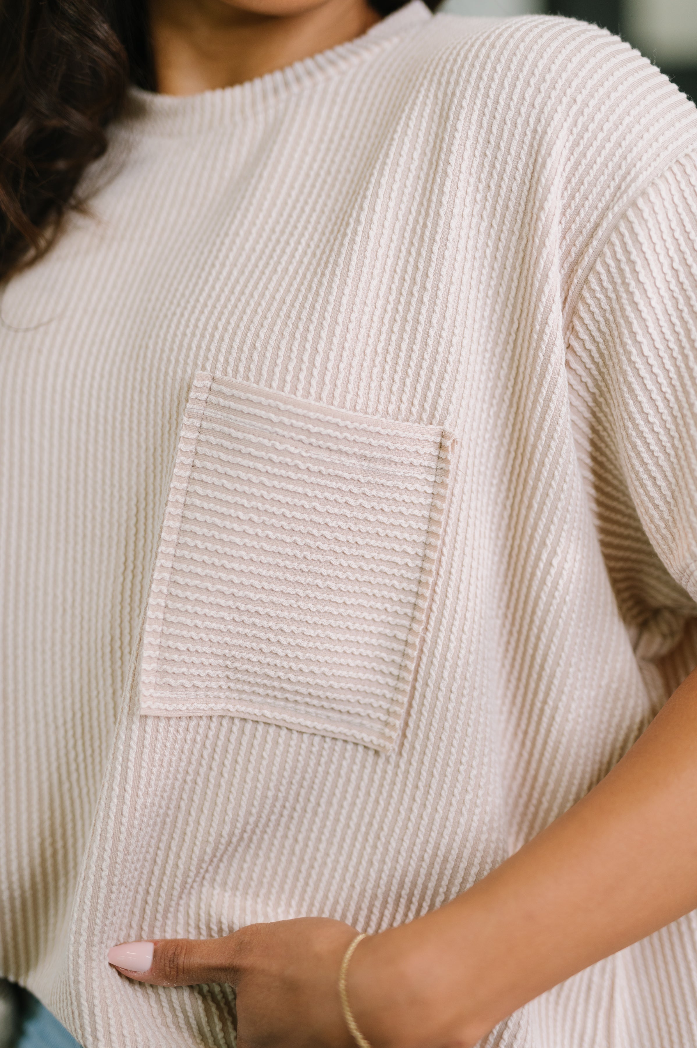 Close-up of ash mocha textured boxy tee with chest pocket, worn by person with hand resting near waist and bracelet visible