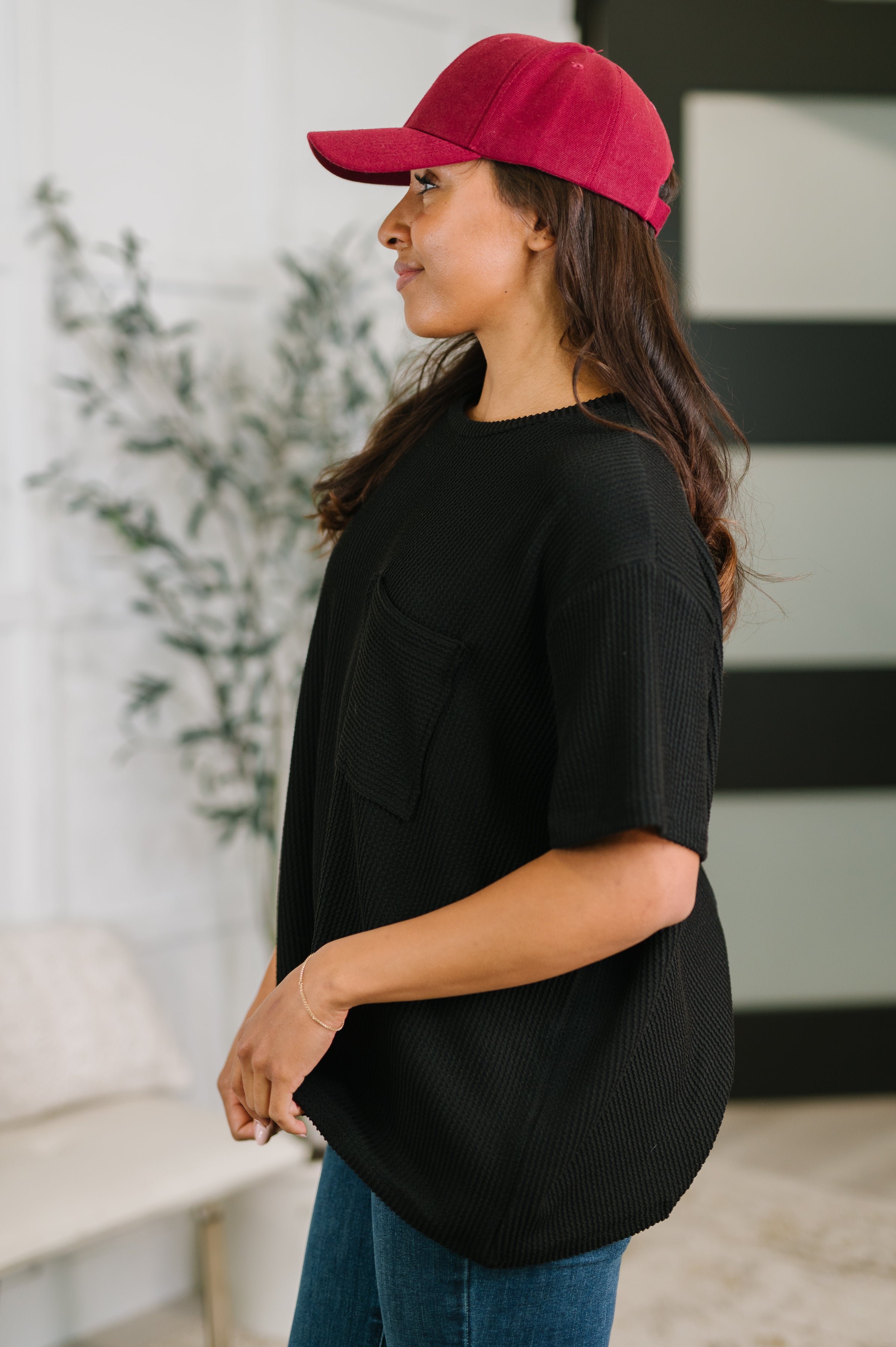 Profile of woman wearing black textured boxy tee with chest pocket and red cap standing indoors near plant and window
