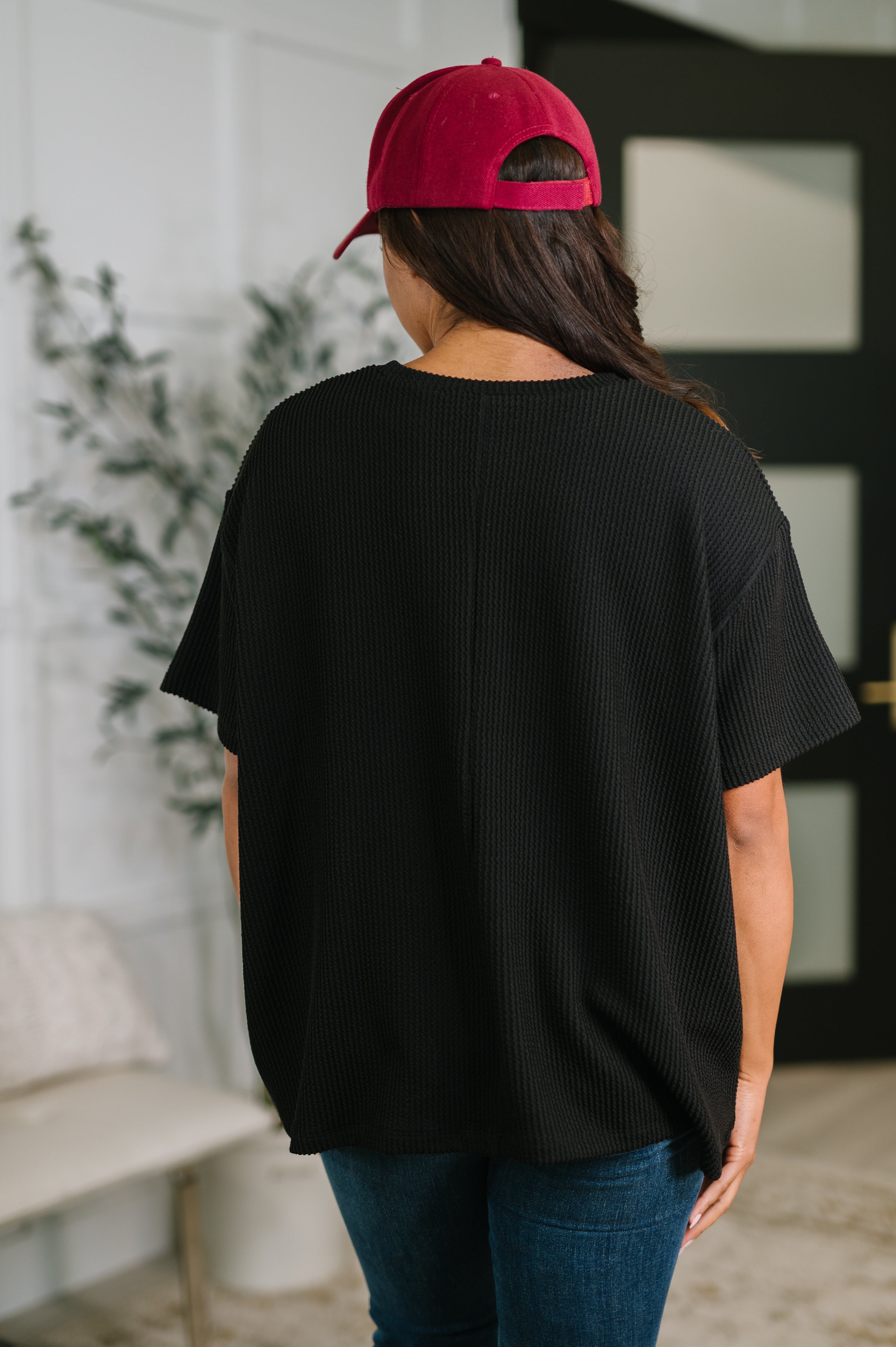 Woman wearing a black textured boxy tee with short sleeves and a red cap indoors with blurred background elements