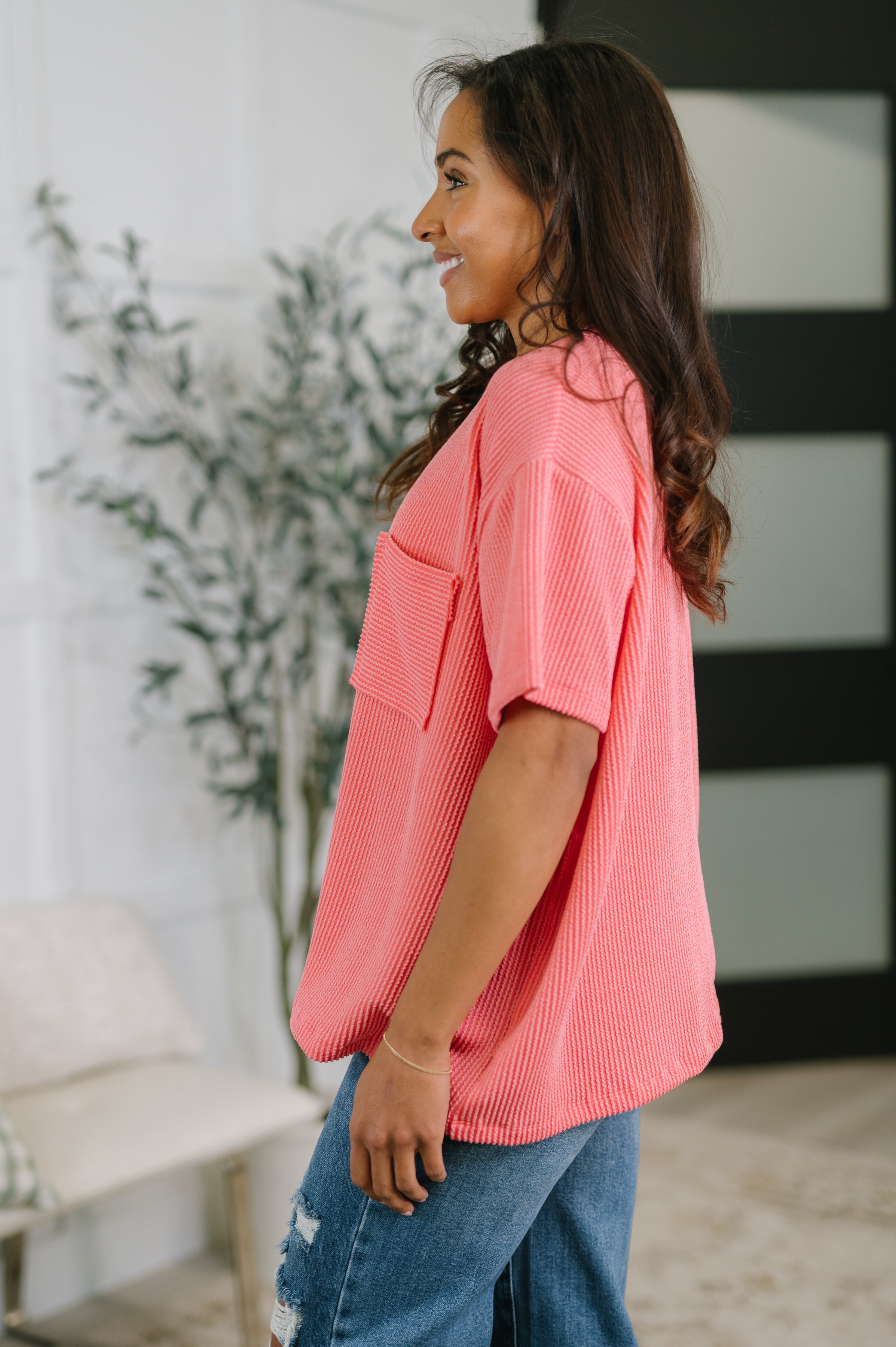 Side view of woman wearing salmon textured boxy tee with large front pocket paired with ripped blue jeans indoors