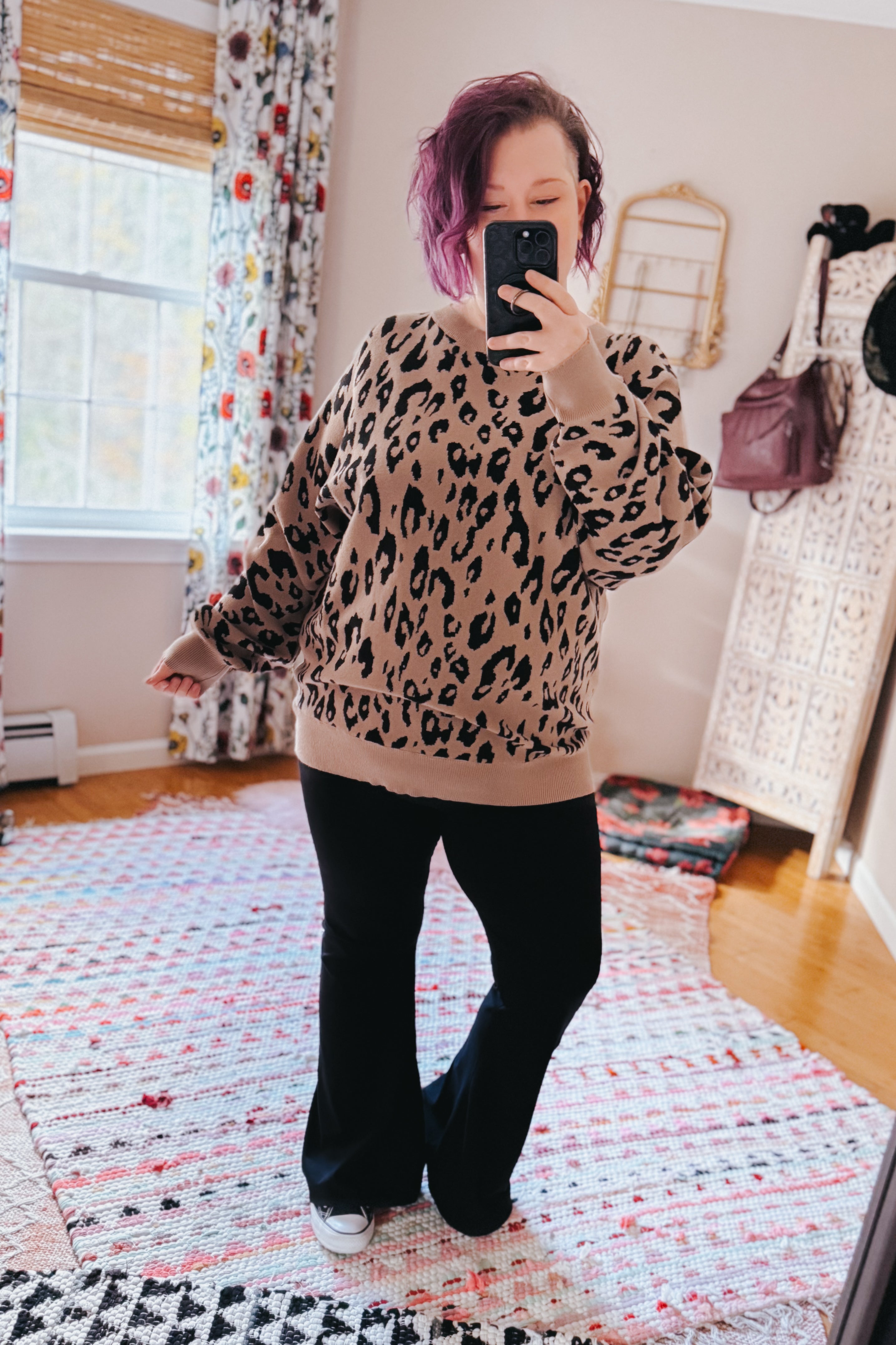 Woman taking a mirror selfie wearing a relaxed mocha sweater with a bold leopard print, paired with black flared pants and sneakers in a bright room.