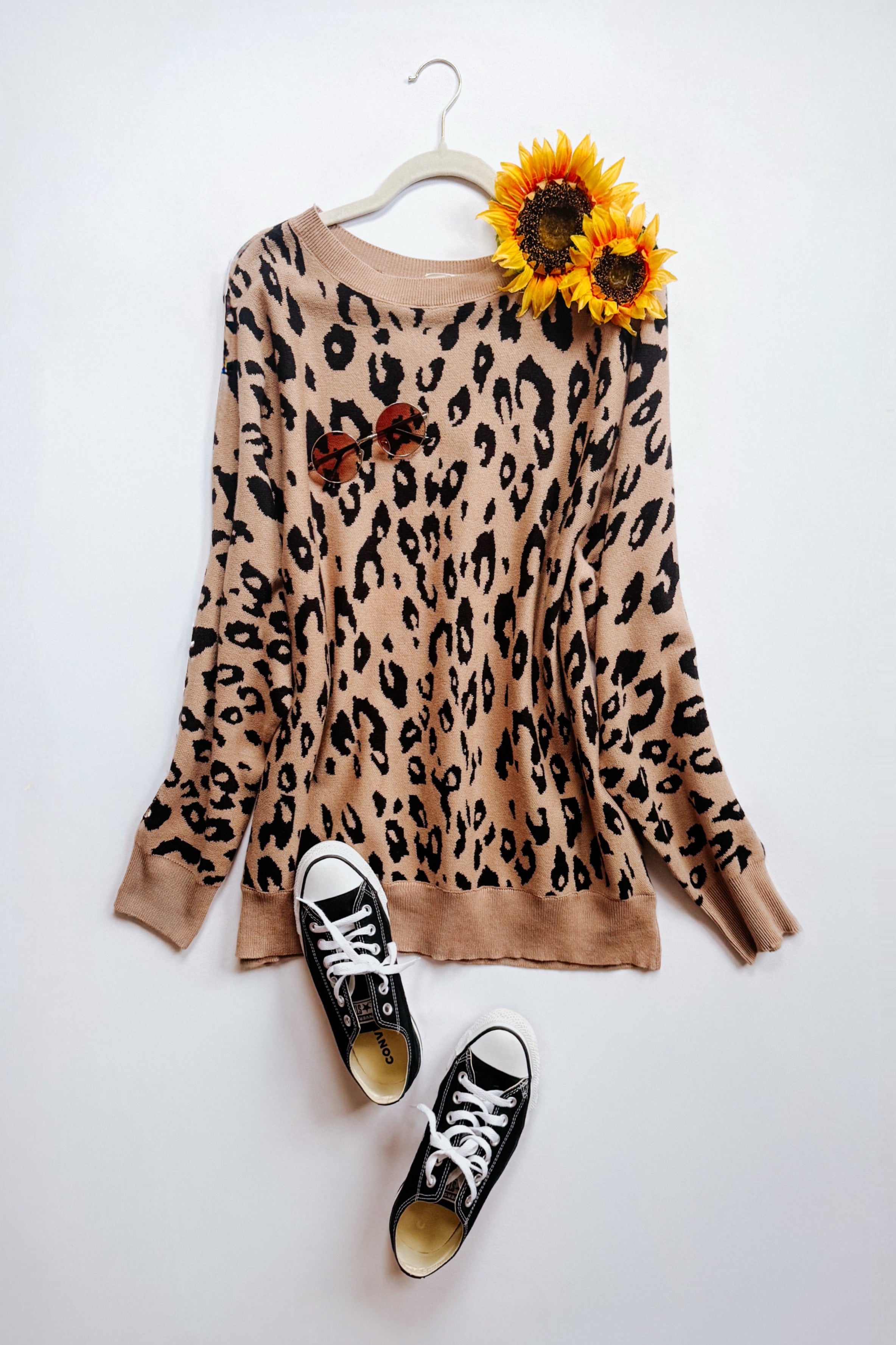 Mocha sweater with a black leopard pattern laid flat, styled with black canvas sneakers, round sunglasses, and two sunflowers on top.