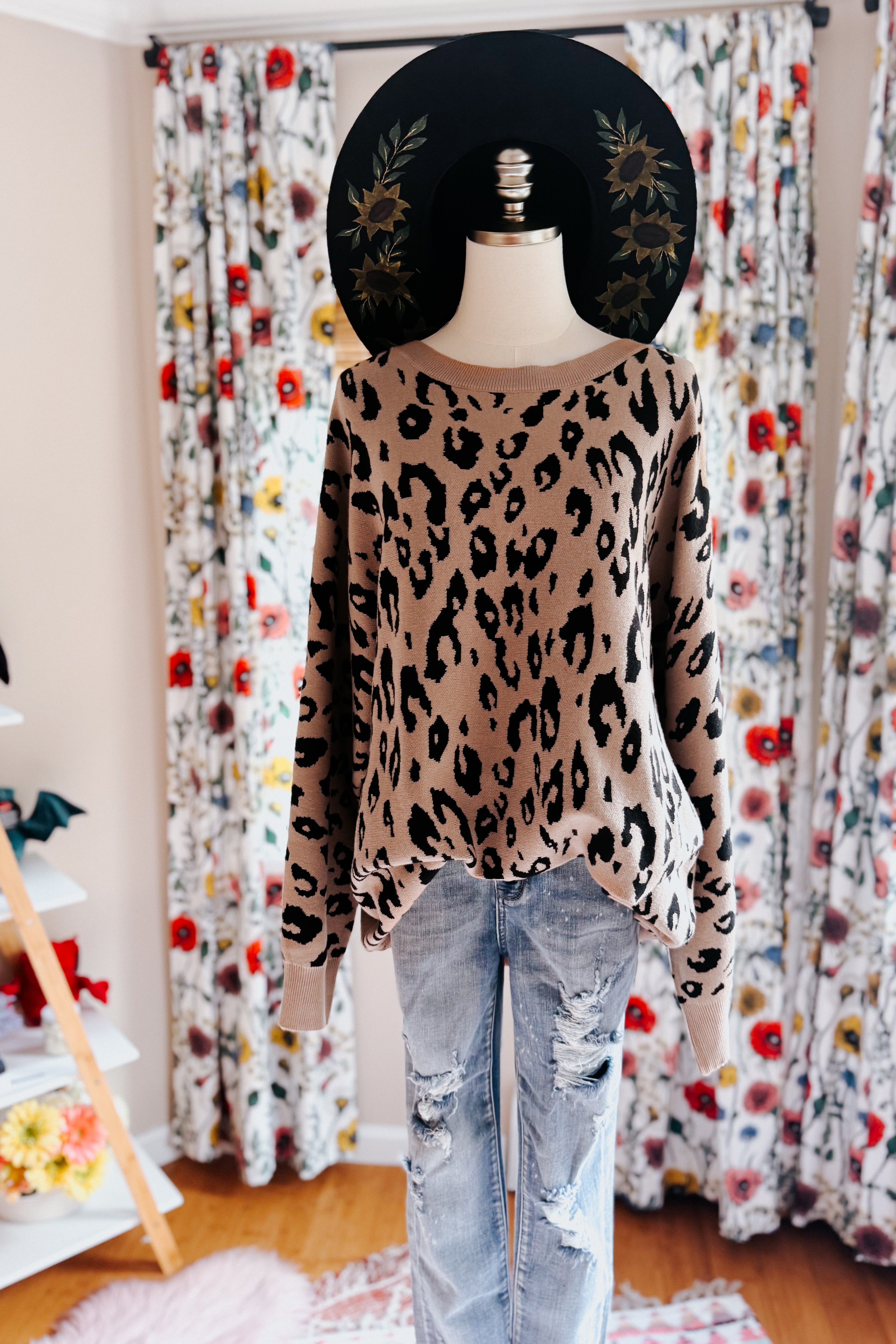Mannequin dressed in a mocha sweater with a black leopard pattern paired with distressed light blue jeans, styled with a black hat featuring sunflower designs.