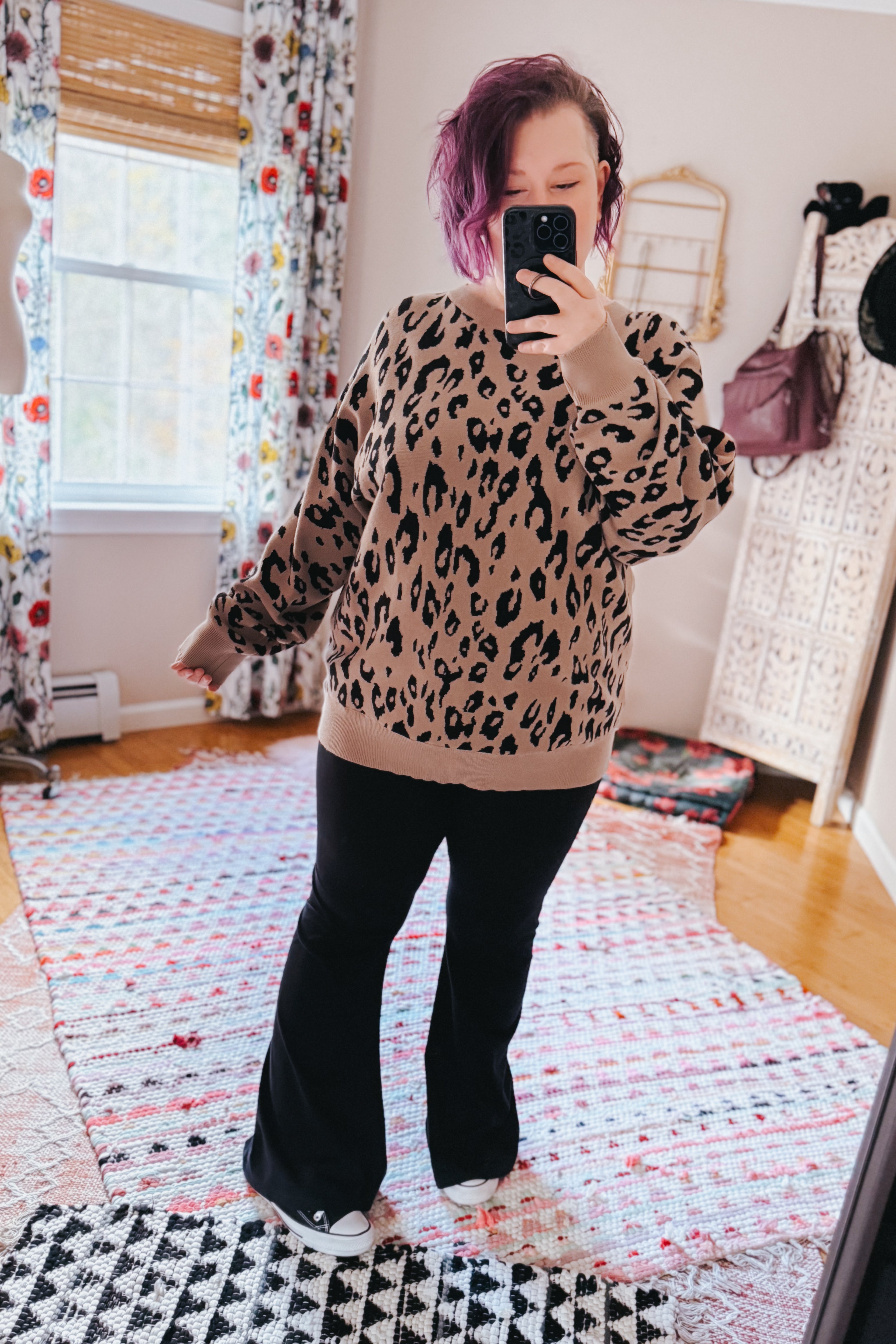 Woman taking a mirror selfie wearing a mocha sweater with a black leopard pattern, paired with black flared pants and sneakers in a bright room.