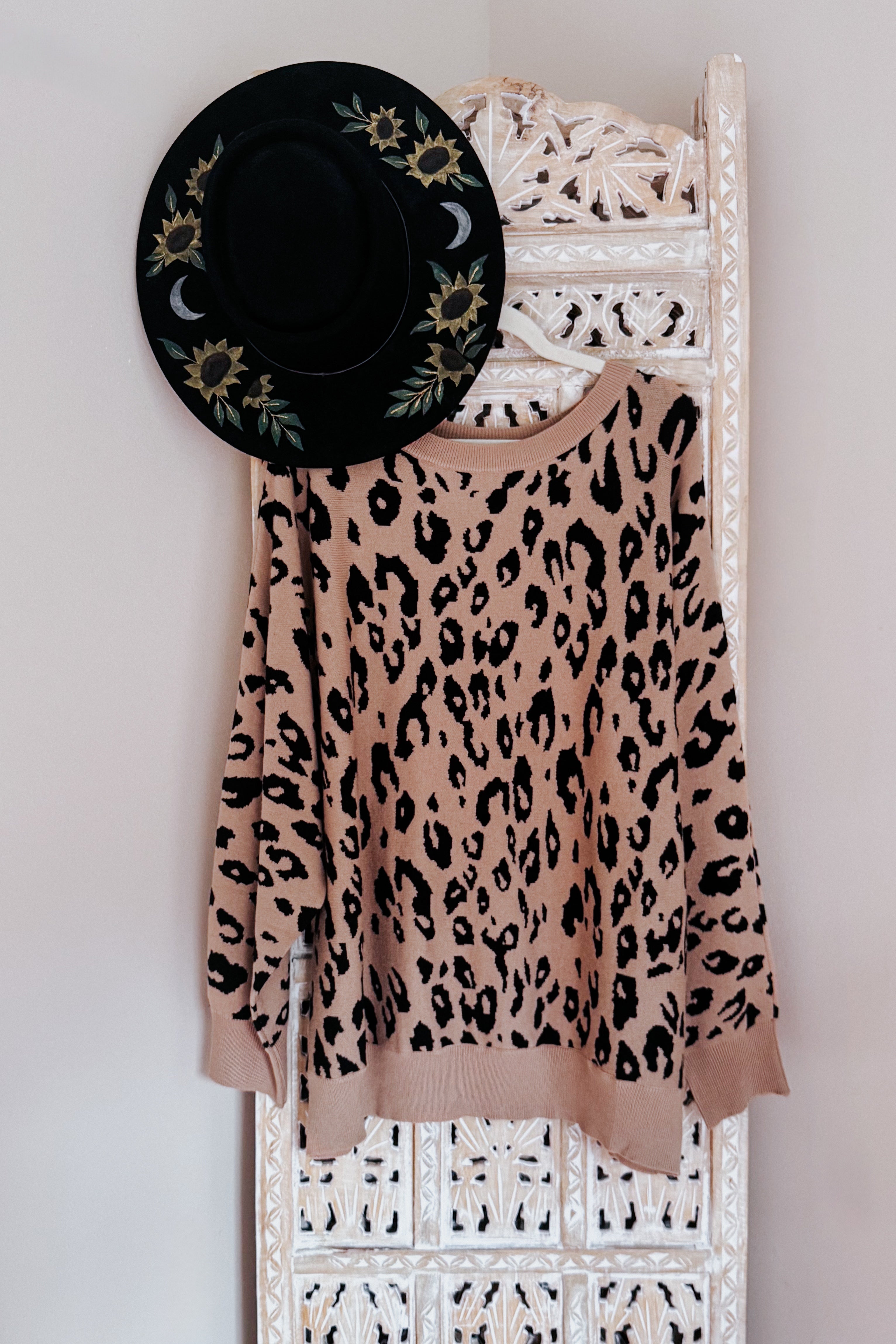 Mocha sweater with a bold black leopard print hanging on a carved white wooden screen next to a black hat decorated with sunflowers and crescent moons.