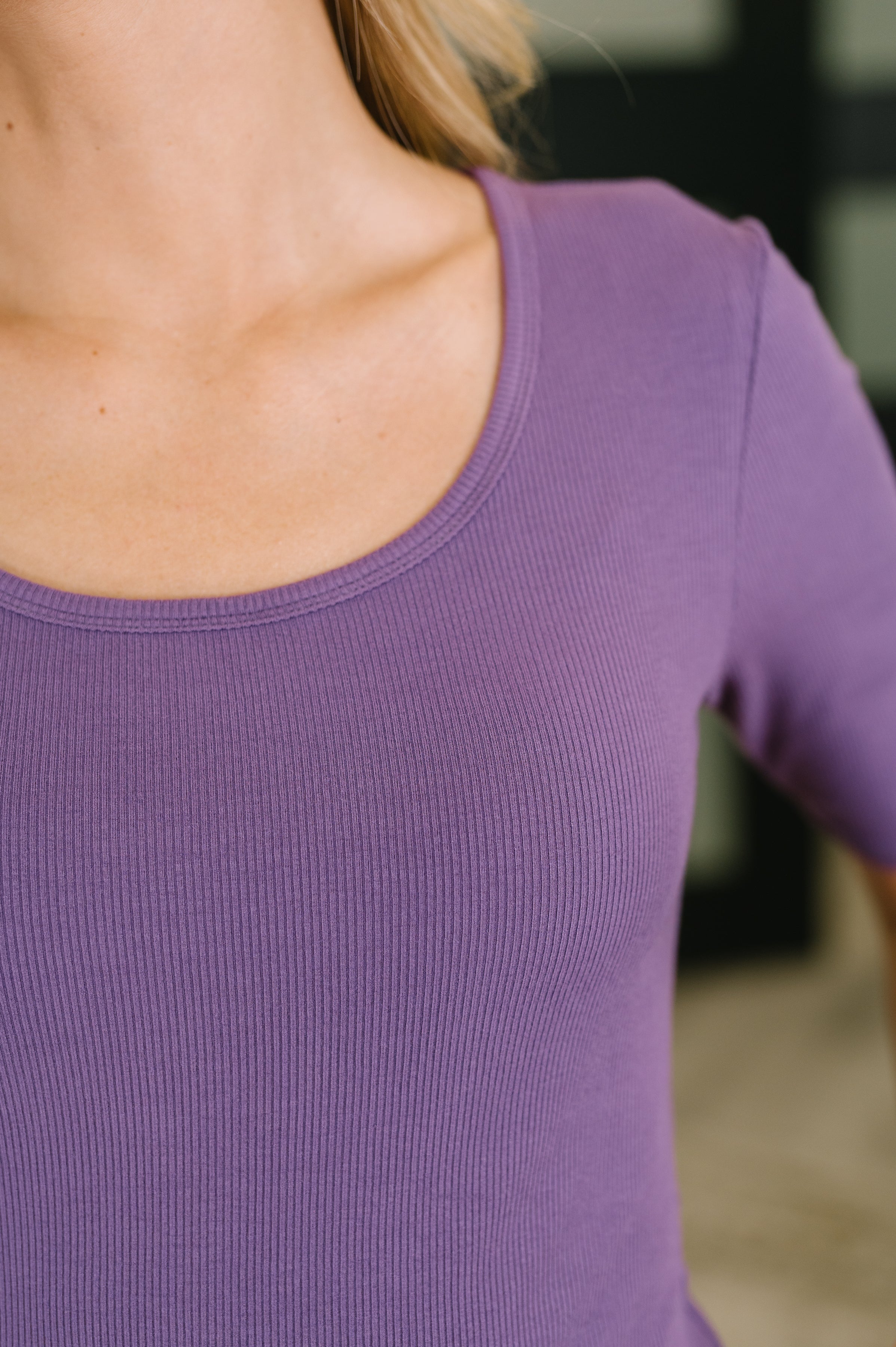 Close view of a woman wearing a purple ribbed knit top with a rounded U neckline and fitted half sleeves