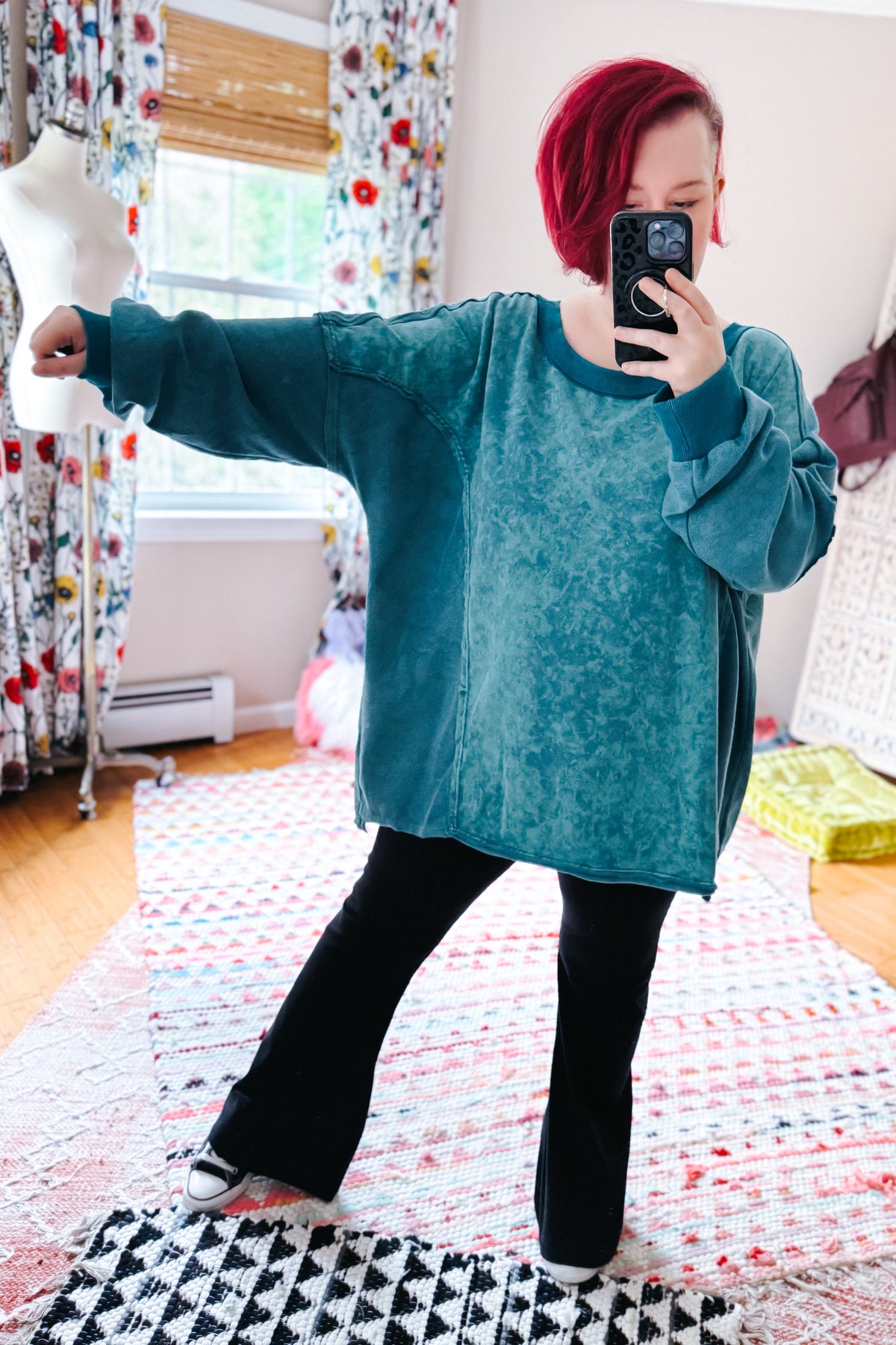 Red haired woman taking a mirror selfie in a blue mineral wash pullover with black flared pants and sneakers on a patterned rug