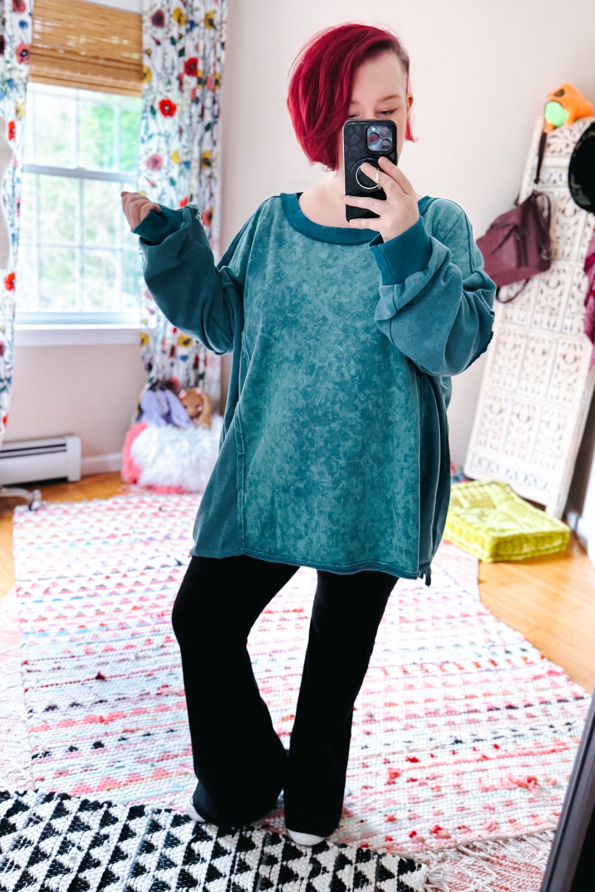 Red haired woman taking a bedroom mirror selfie in a blue mineral wash pullover with oversized sleeves and black pants