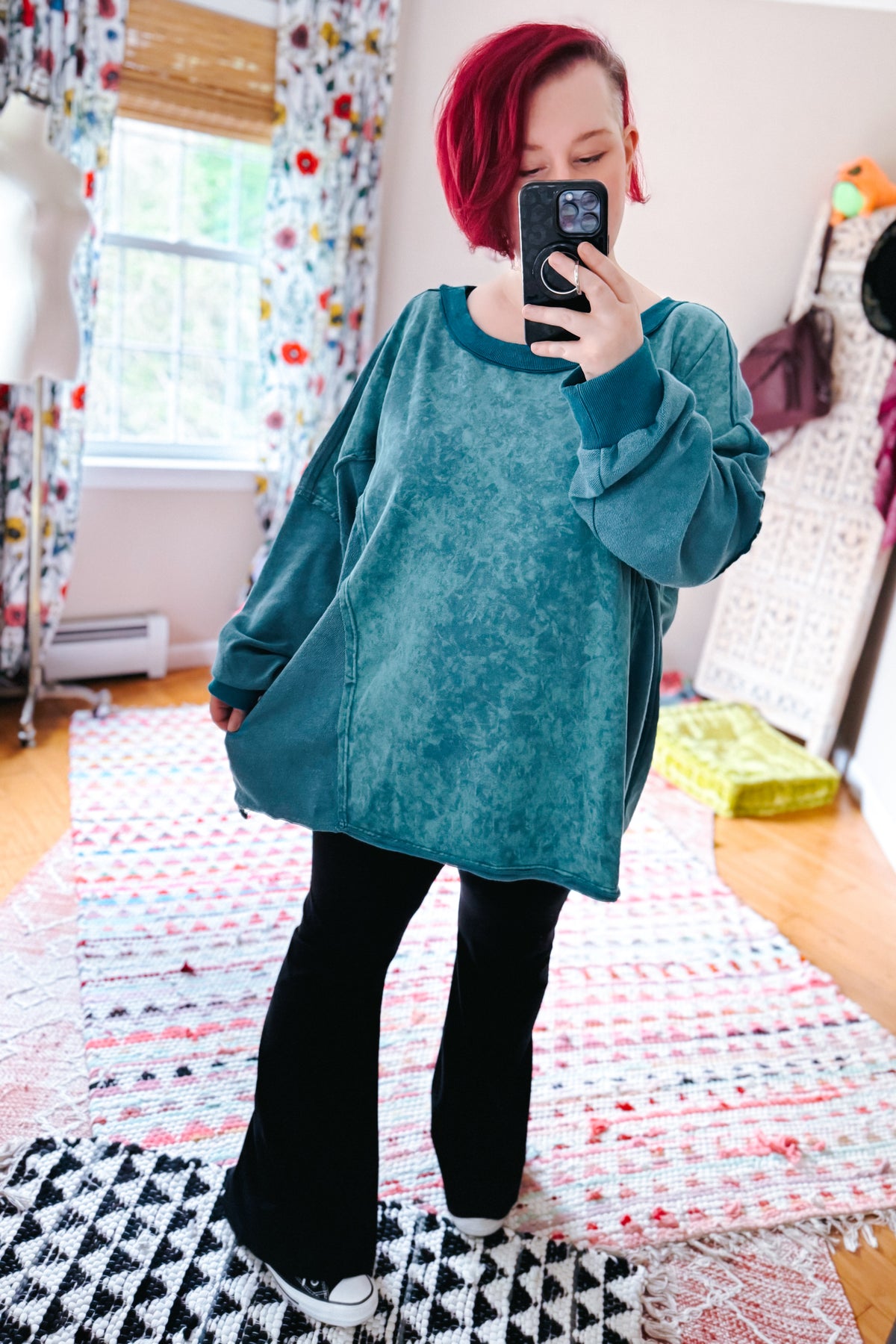 Red haired woman captures a mirror selfie wearing a blue mineral wash pullover, black pants, and sneakers over layered patterned rugs