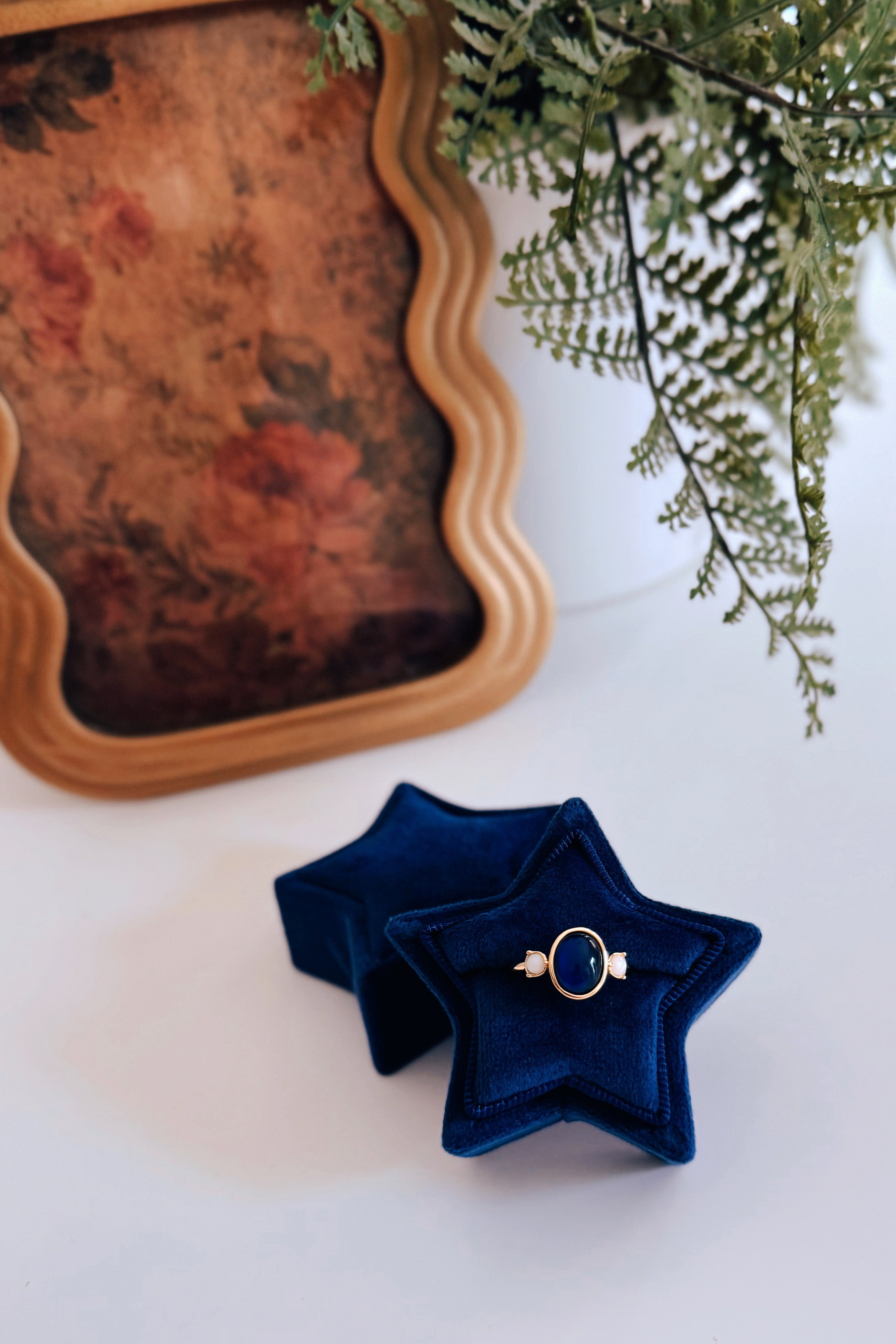 Gold tone ring with dark oval center stone and two small pale round stones displayed on blue star velvet box next to fern leaves