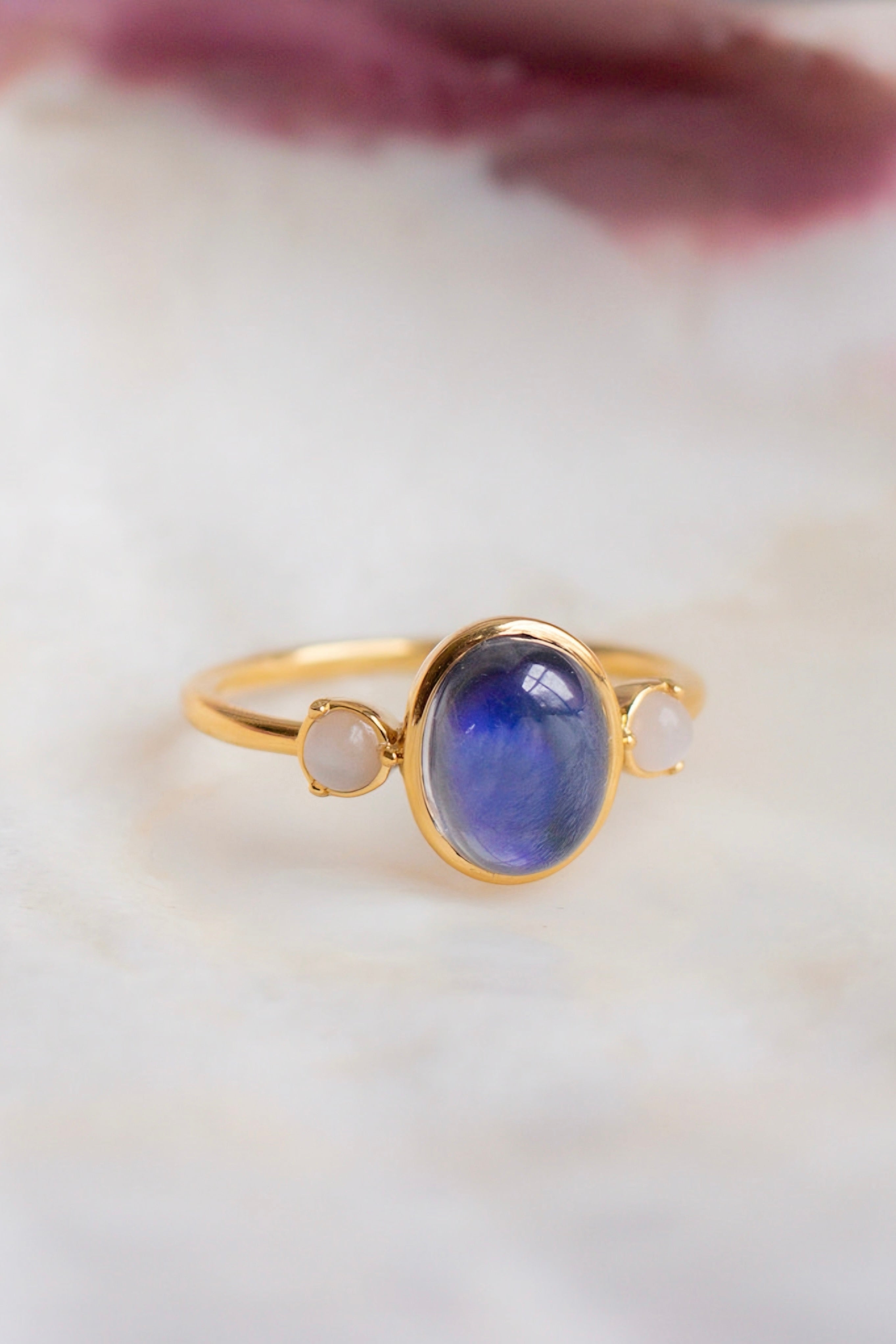 Gold ring with a deep blue oval stone flanked by two small pale pink stones on a soft marble surface
