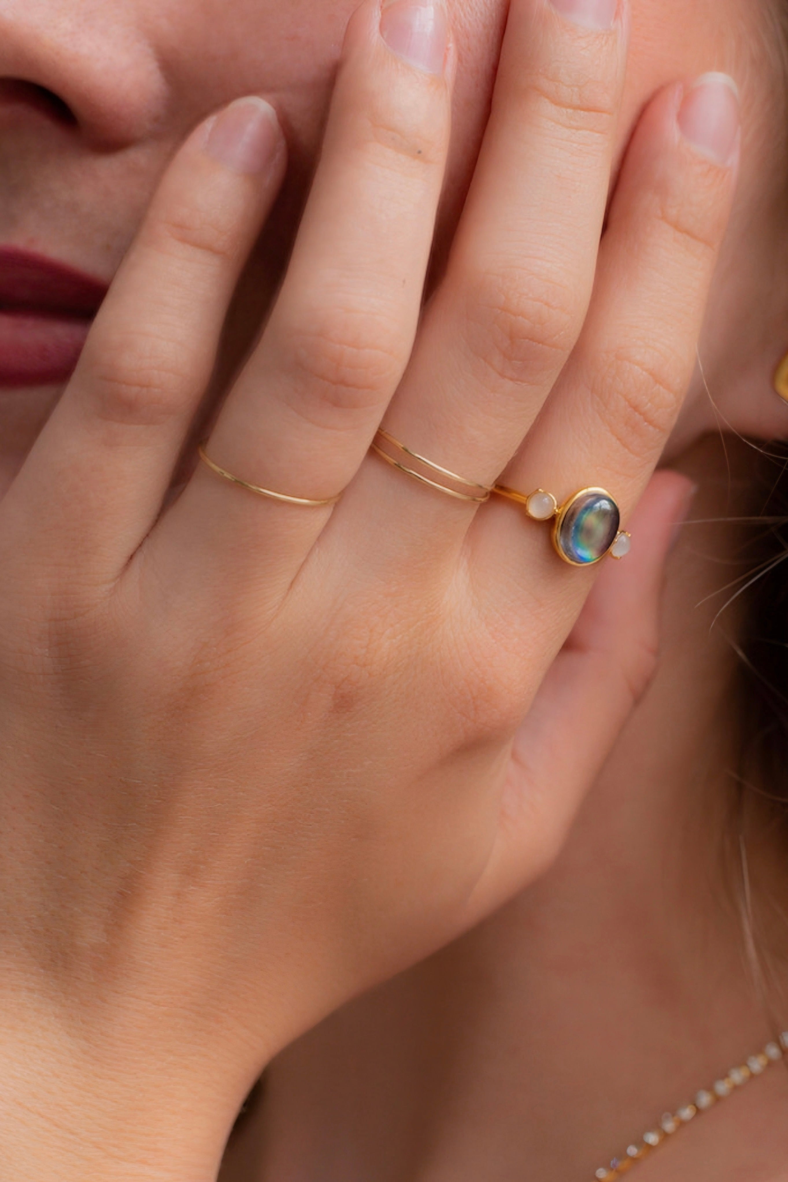 Gold mood ring with iridescent oval stone and two small round stones worn with thin gold bands on hand near face