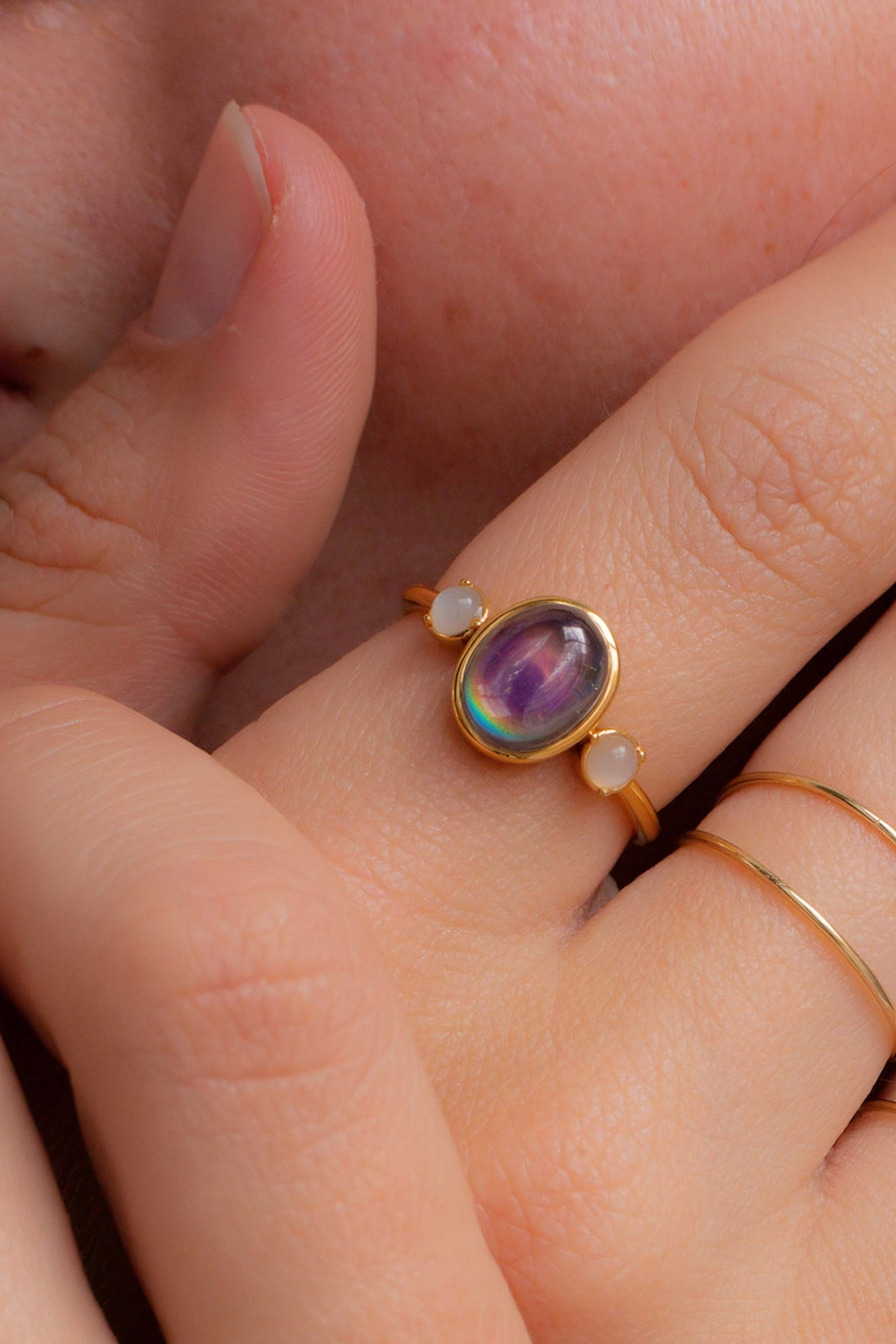 Gold ring with oval iridescent purple stone flanked by two small round white stones on finger