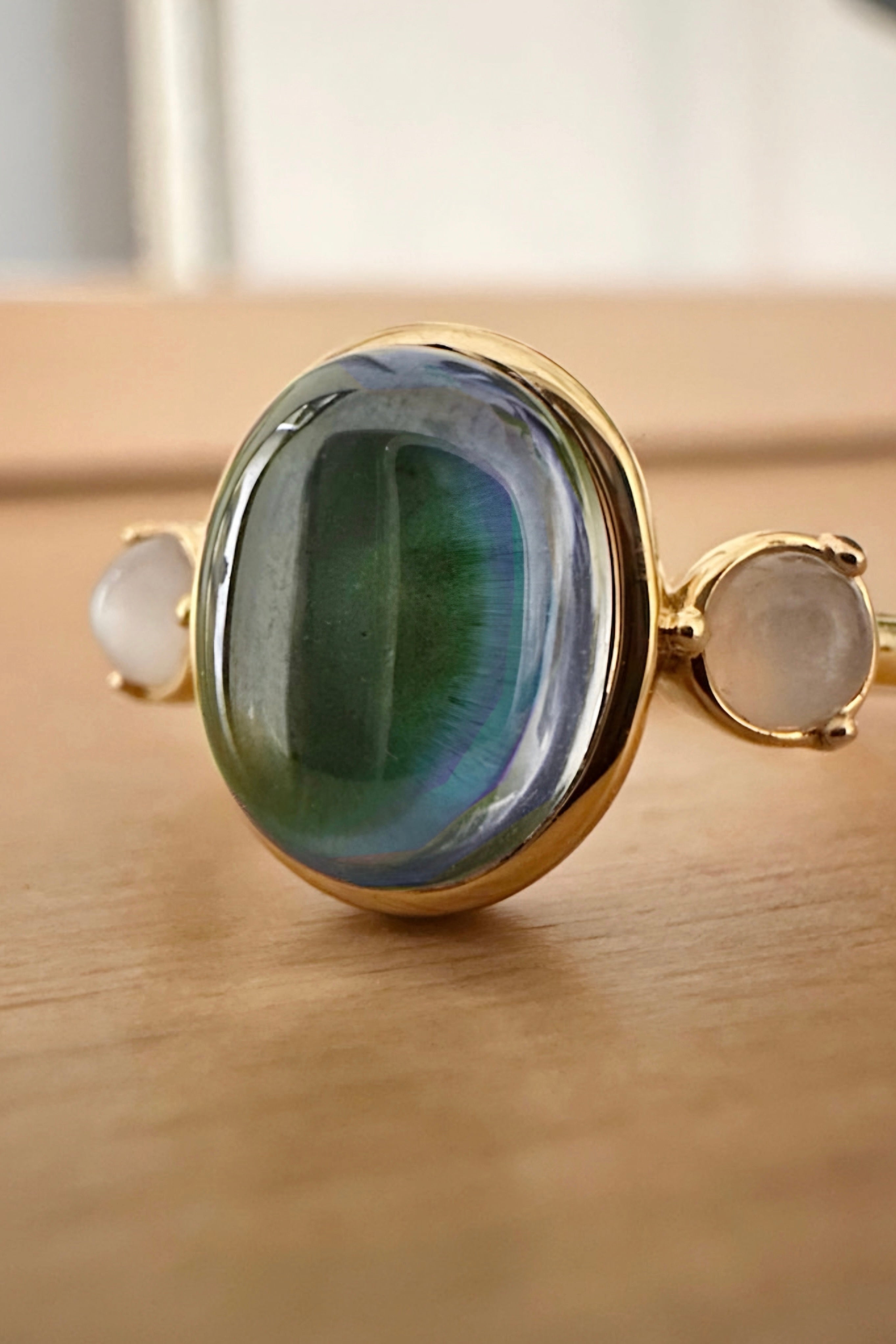 Gold band mood ring with a large iridescent blue-green stone and two small pale round stones on wood surface