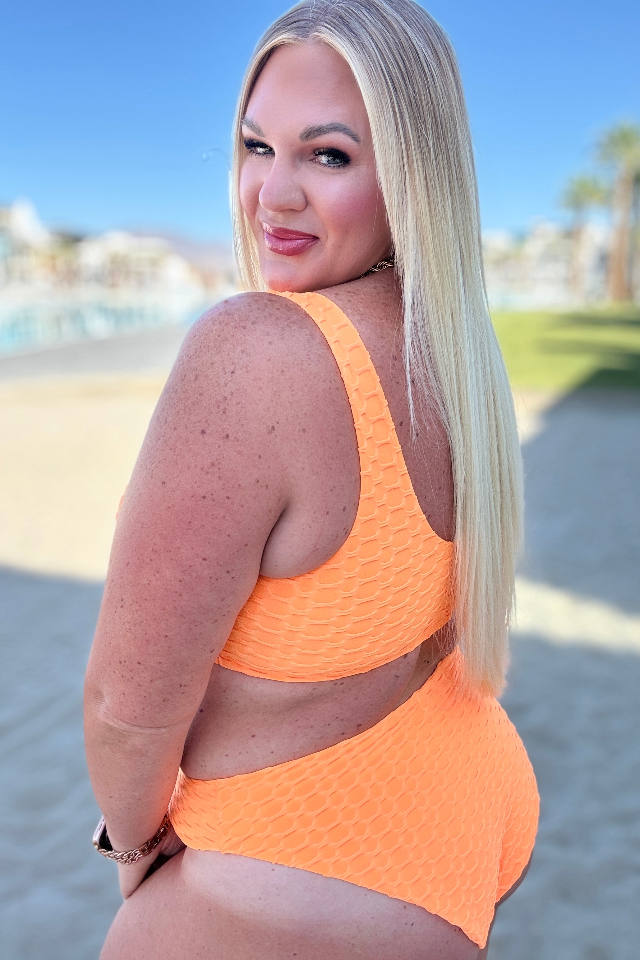 Plus-size woman with long blonde hair wearing vibrant textured orange swimwear outdoors near waterfront on sunny day