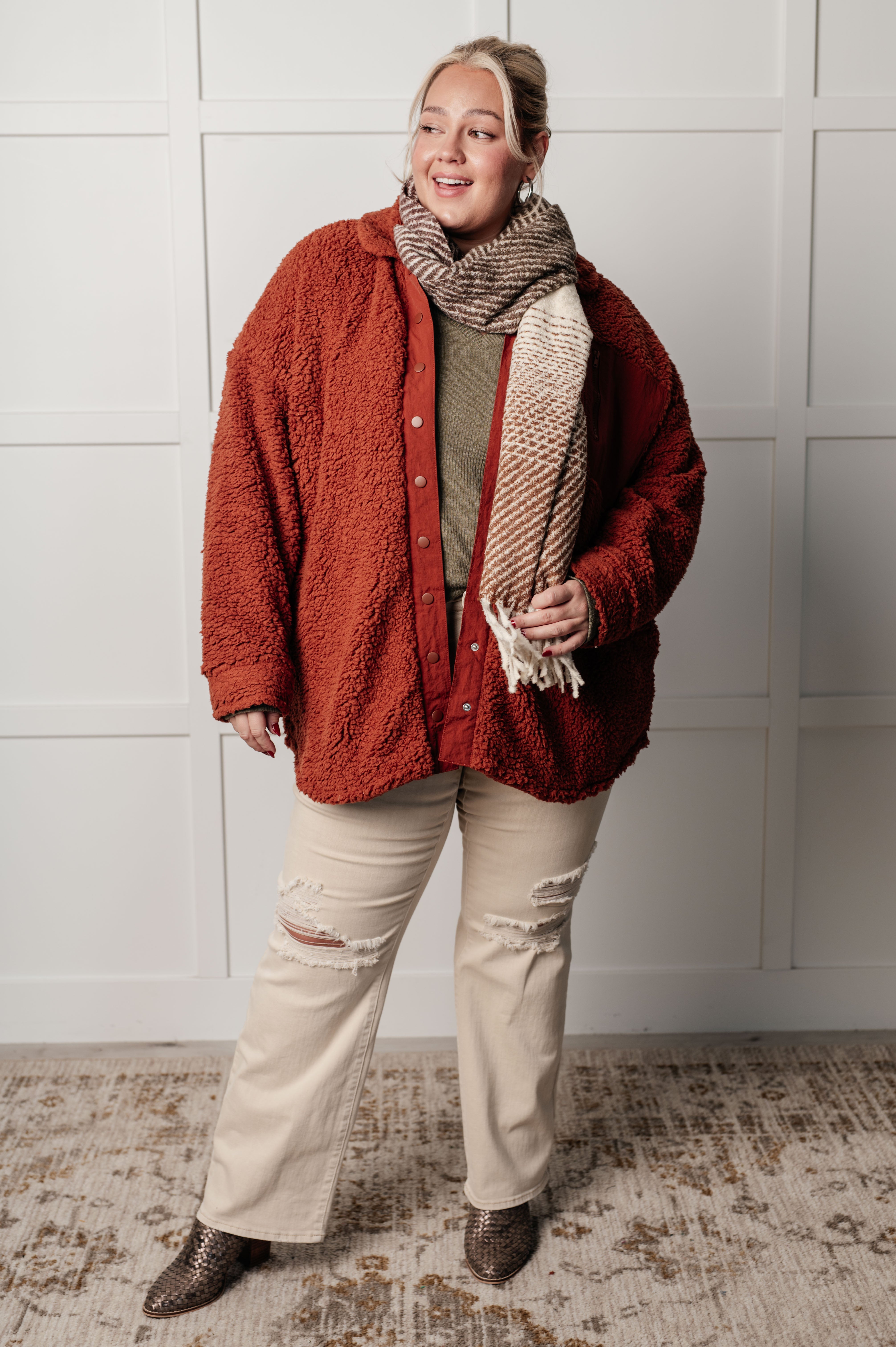 Plus-size model wearing rust textured sherpa jacket with knit scarf, olive ribbed top, distressed light pants, and brown boots indoors