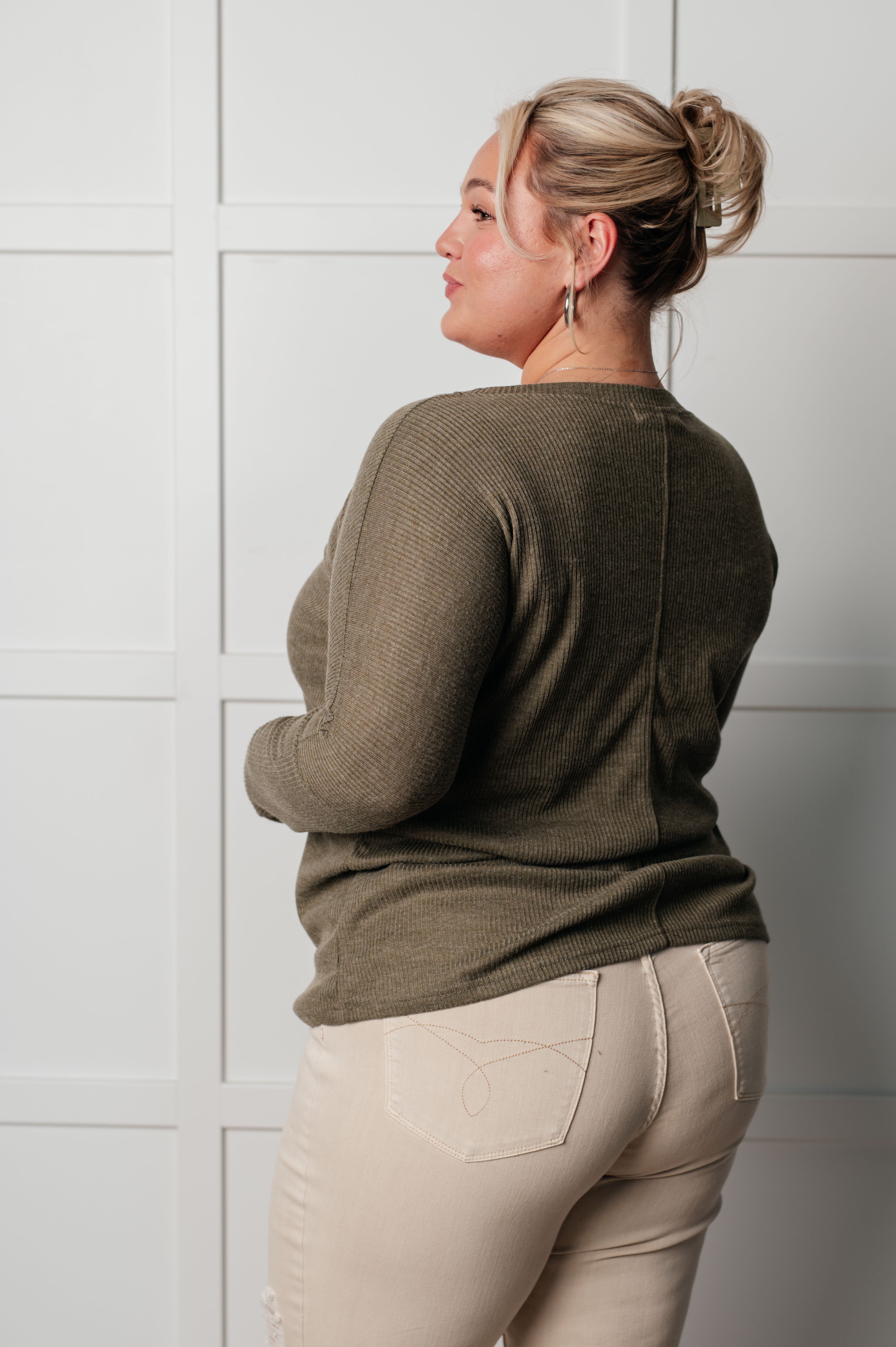 Plus-size model wearing olive ribbed knit top with long sleeves and beige pants against white paneled wall