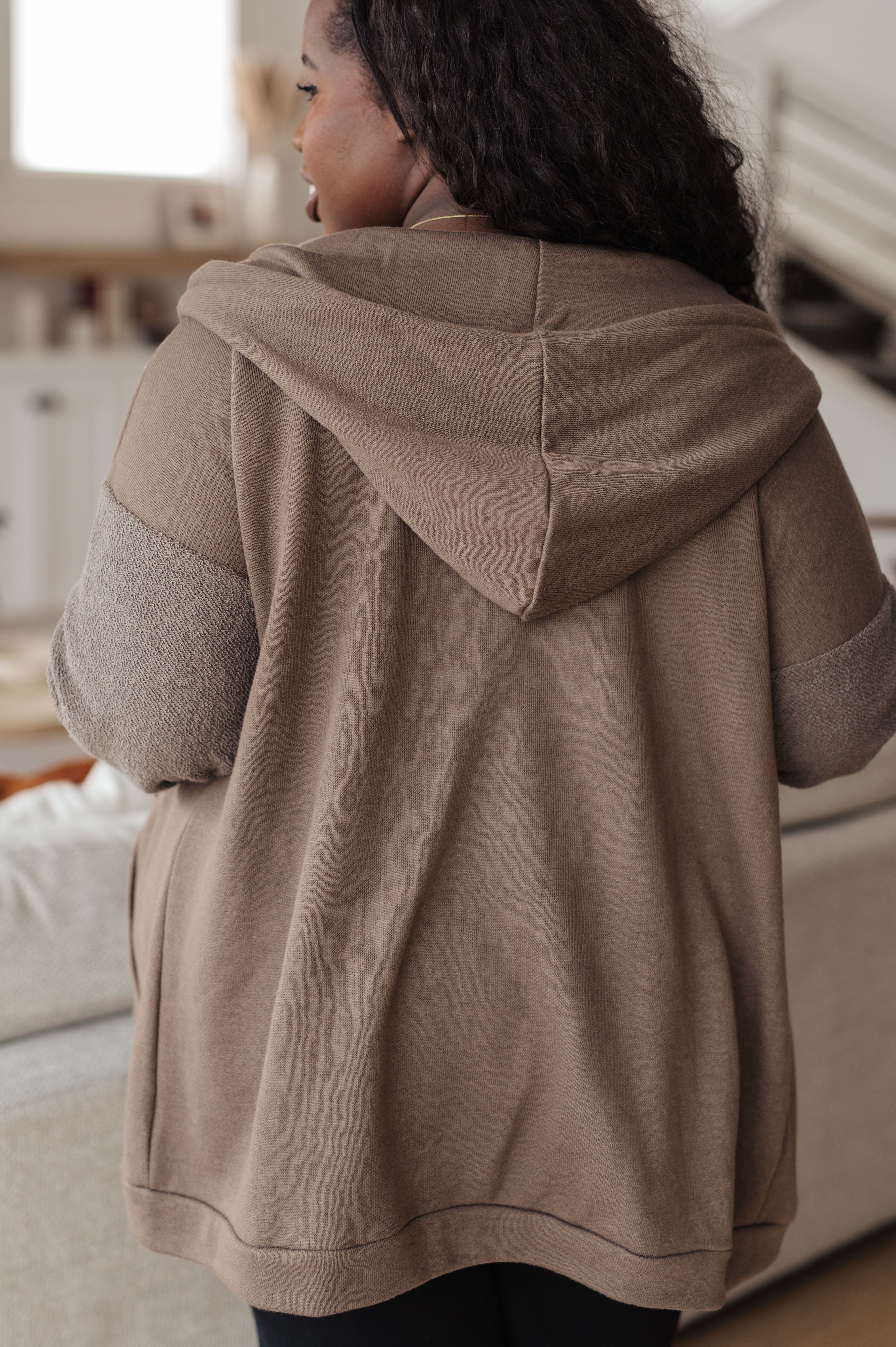 Woman with her back facing the camera wearing a brown zip-up hoodie over over black jeans in a casual indoor setting.