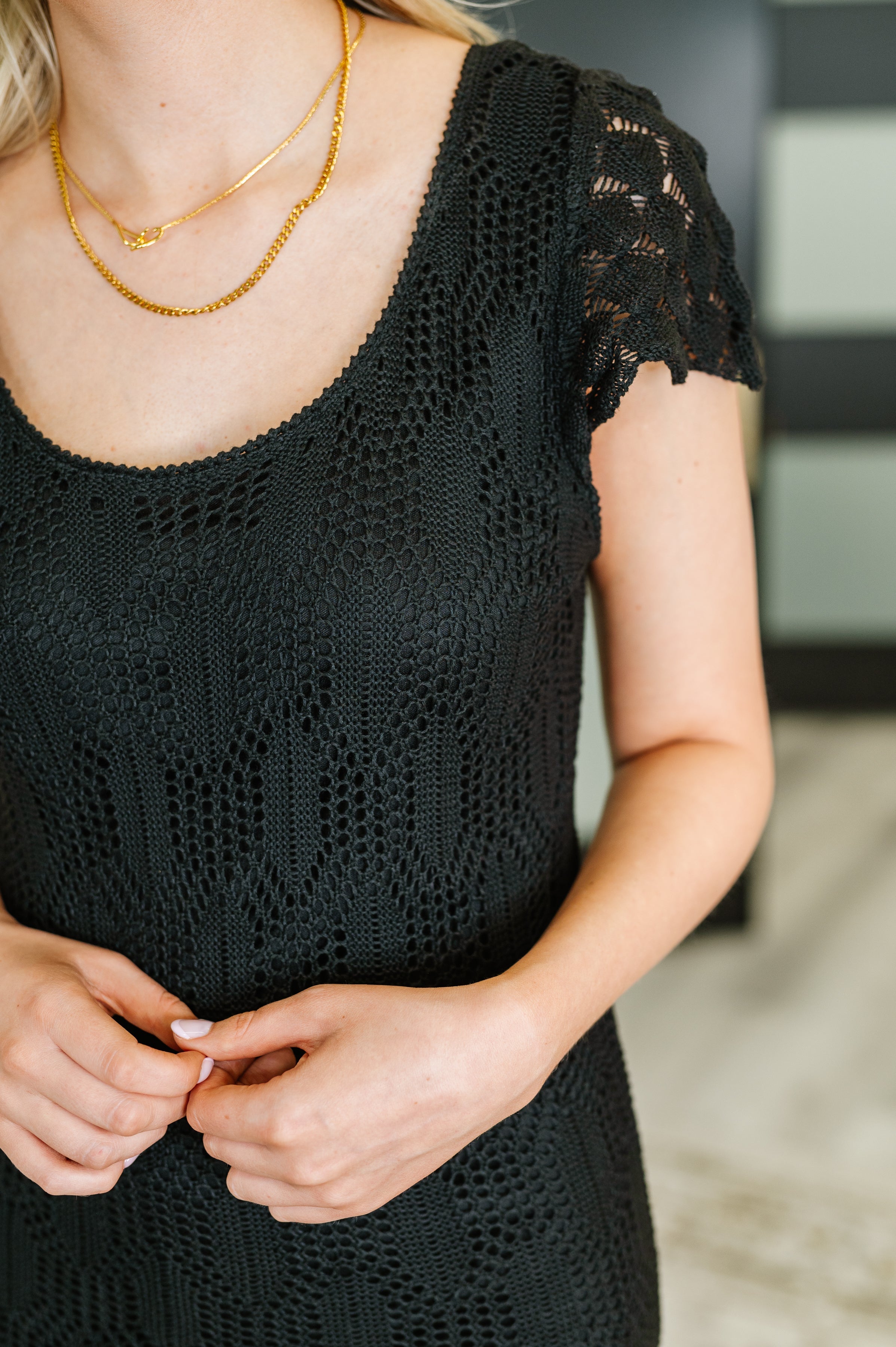 Close-up of woman wearing black lace scoop neck dress with cap sleeves and layered gold necklaces indoors