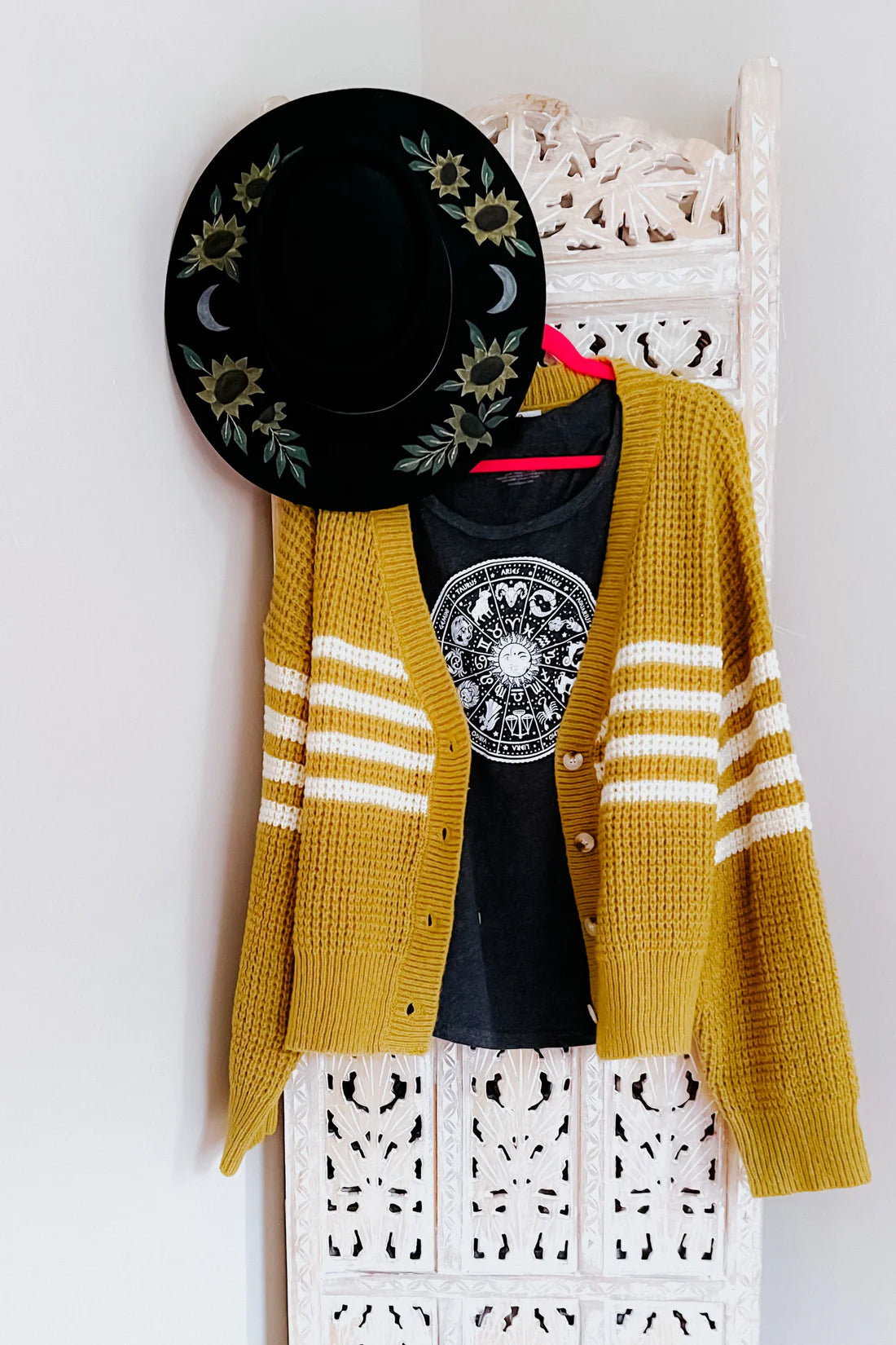 Mustard striped knit cardigan layered over black graphic tee, hanging on carved white panel with black floral hat above